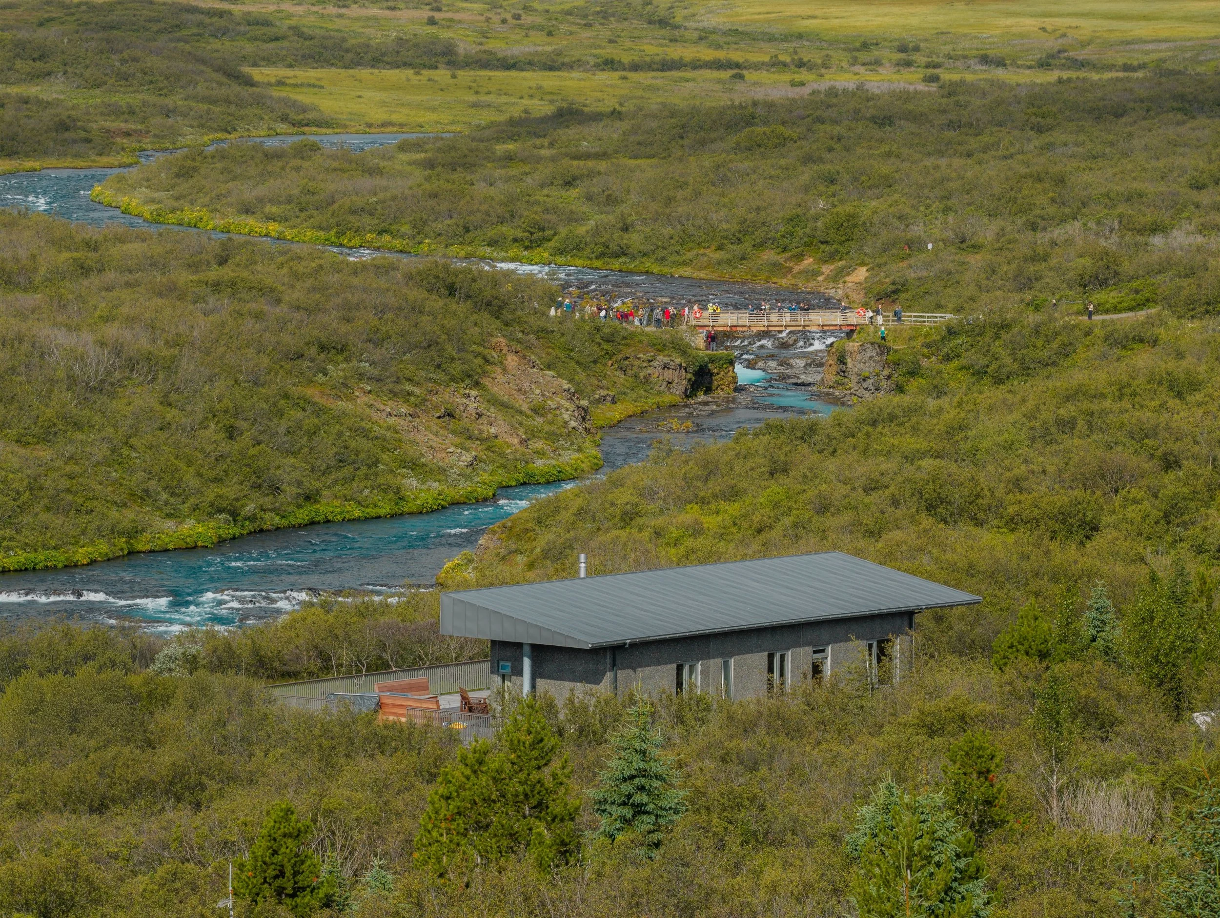 A river flowing through a lush green landscape with forests and grassy fields. A bridge with people on it crosses the river, and there is a building with a metal roof in the foreground.