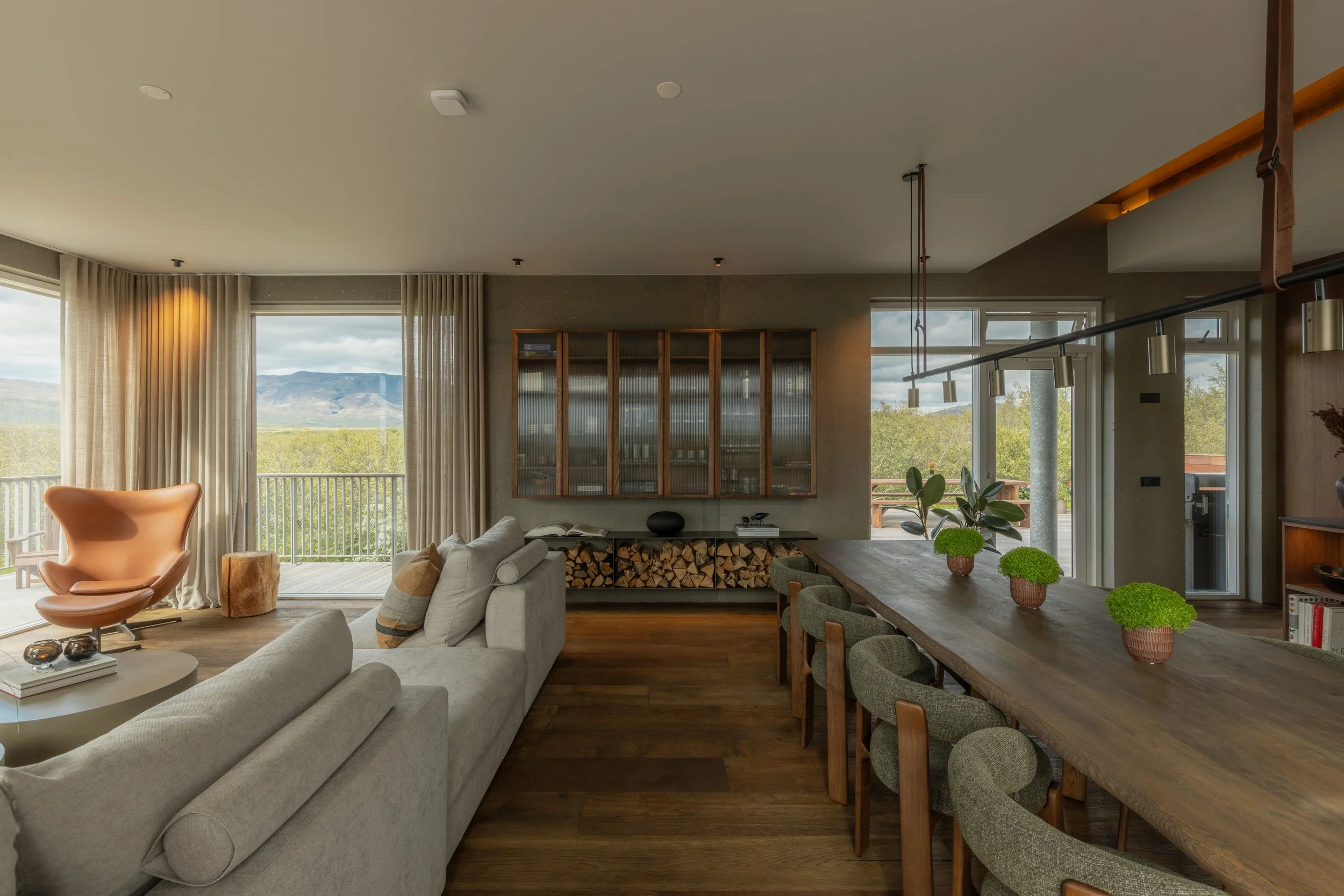Living room with large windows, a beige sofa with striped pillows, a round coffee table, modern chairs, a wooden dining table with green potted plants, and a wall cabinet filled with books, overlooking a scenic mountain view.