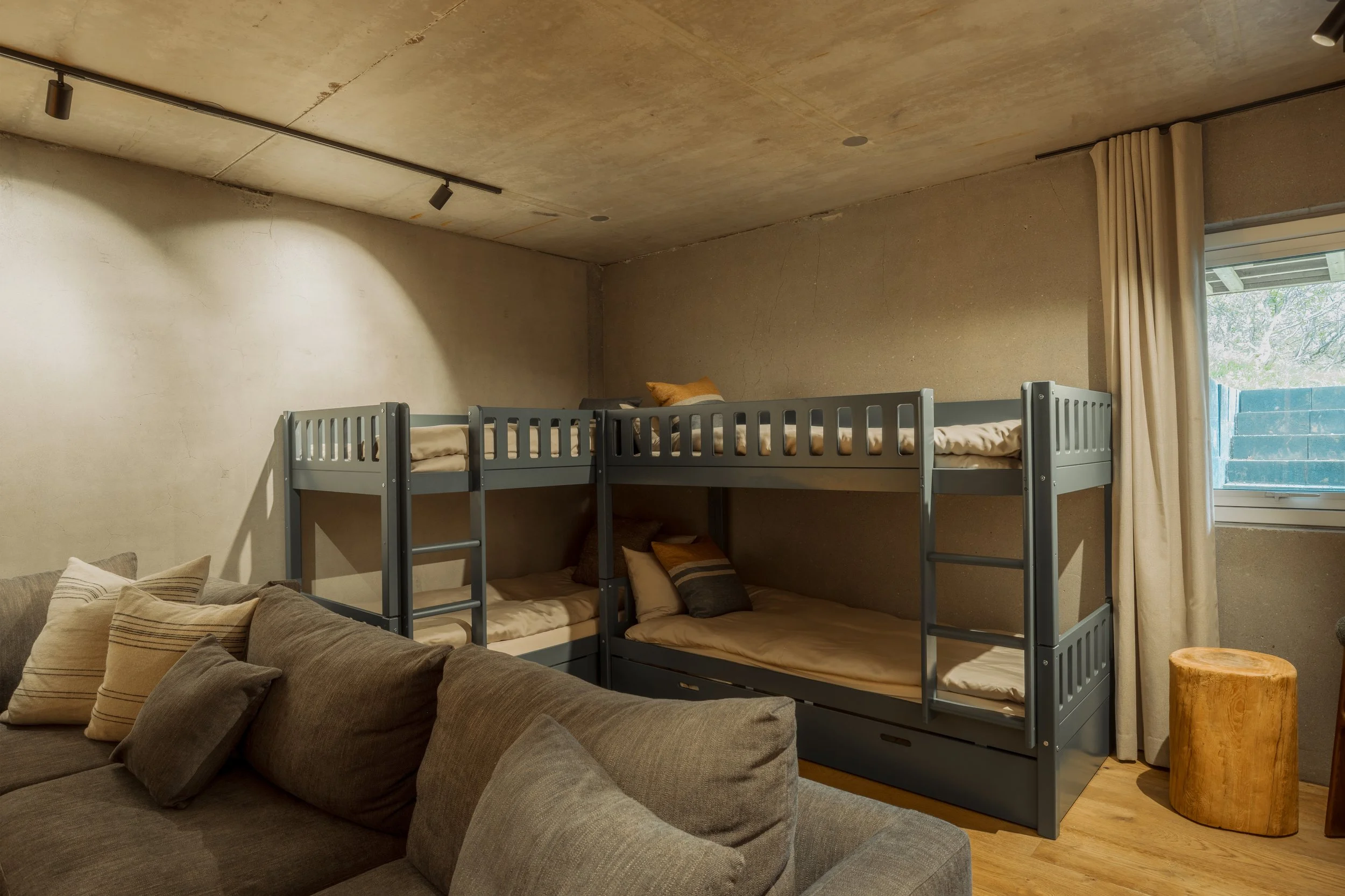 A room with concrete walls, a window with beige curtains, and a bunk bed with beige bedding. There's a gray sofa with striped pillows in the foreground and a small wooden stool.