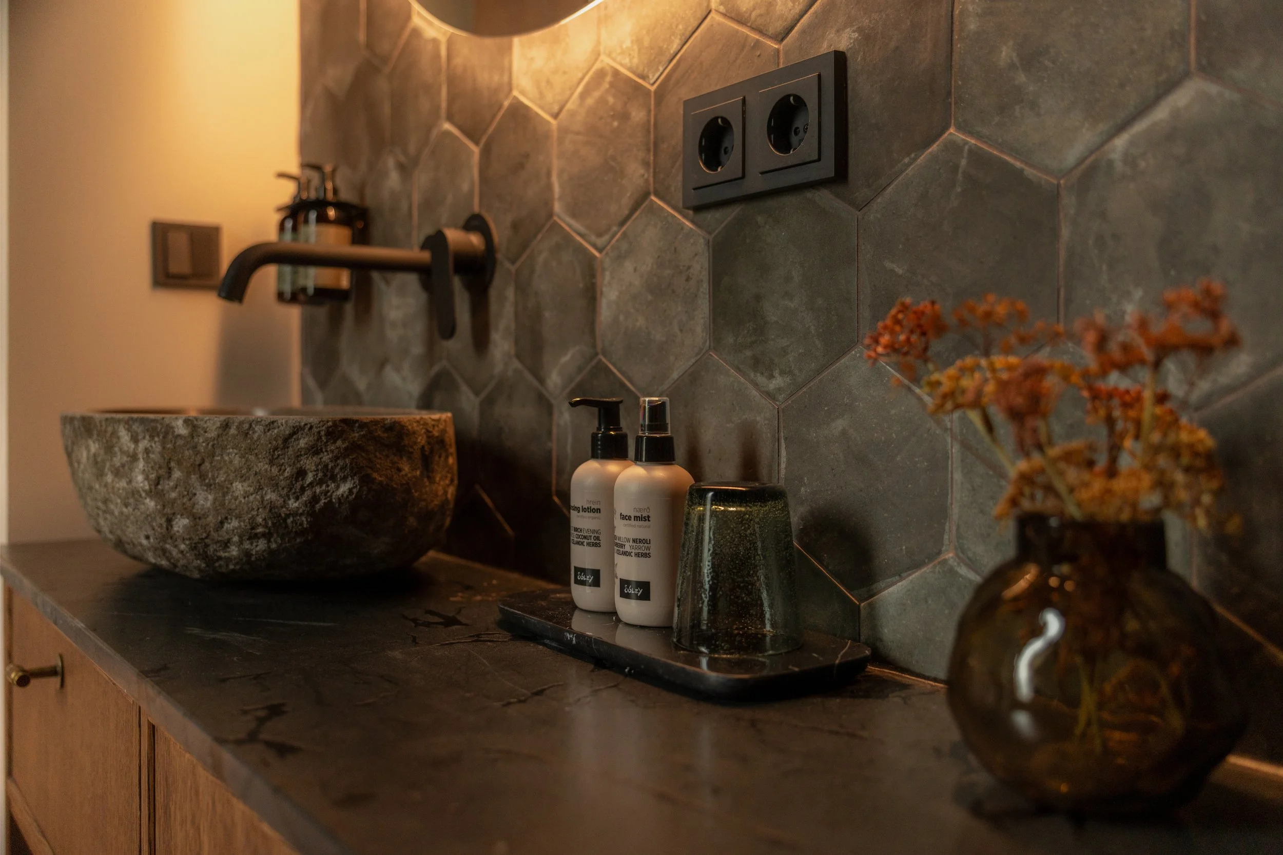 A bathroom vanity with a round stone sink, a black faucet, and two bottles of skincare products on a black tray, against a dark gray tiled wall with electrical outlets.