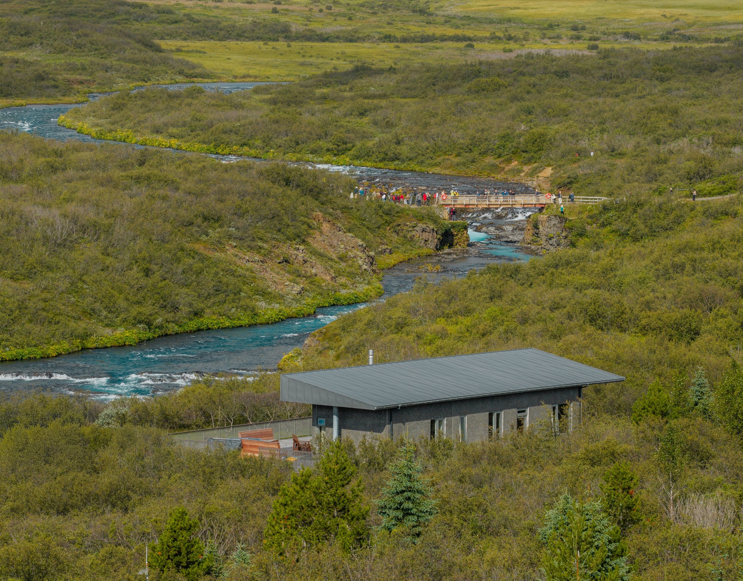 A scenic view of a river flowing through green hills, with a modern building with a metal roof and outdoor seating in the foreground.