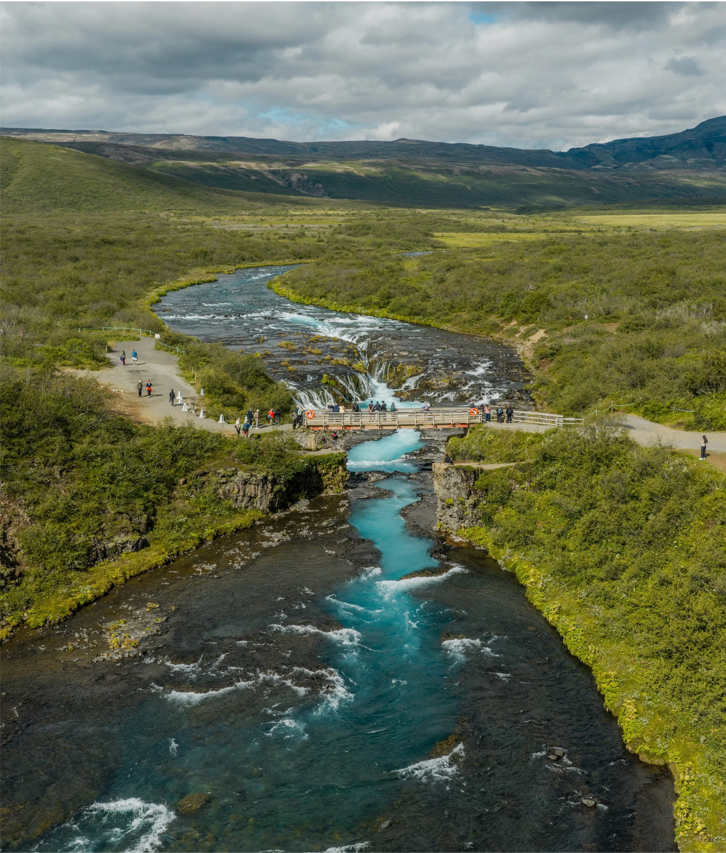 A scenic landscape featuring a river flowing through a lush green valley with hills and mountains in the background under a cloudy sky.