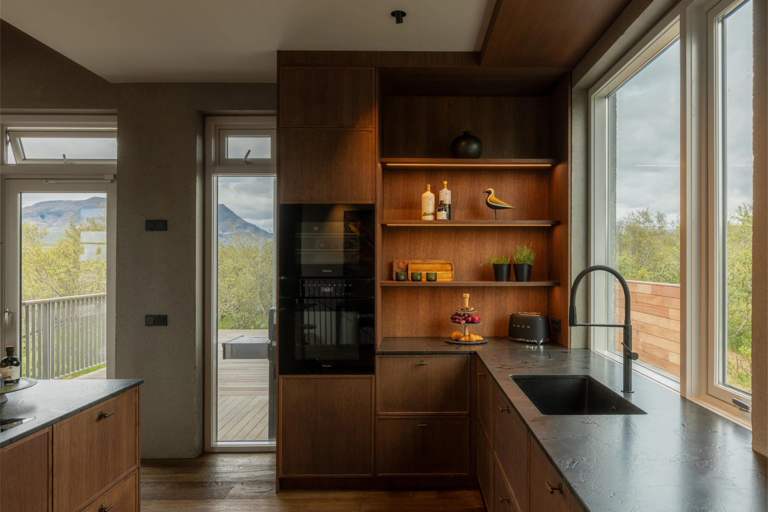 A modern kitchen with wood cabinetry, large windows showing mountain views, and a sliding door leading to an outdoor deck. The kitchen has a black countertop, a black faucet, and shelves with decorative items, plants, and bottles.