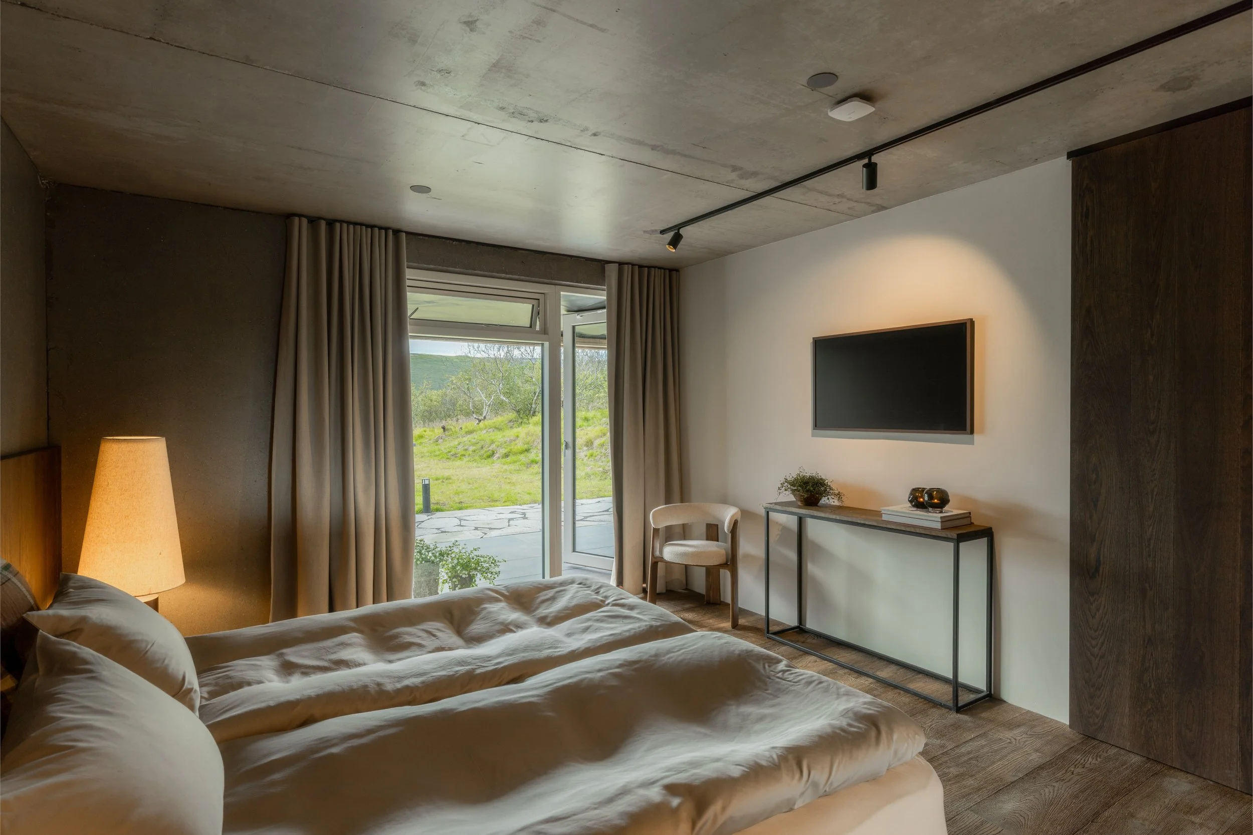 Modern bedroom with bed, nightstand, and lamp, large window with beige curtains showing outdoor greenery.