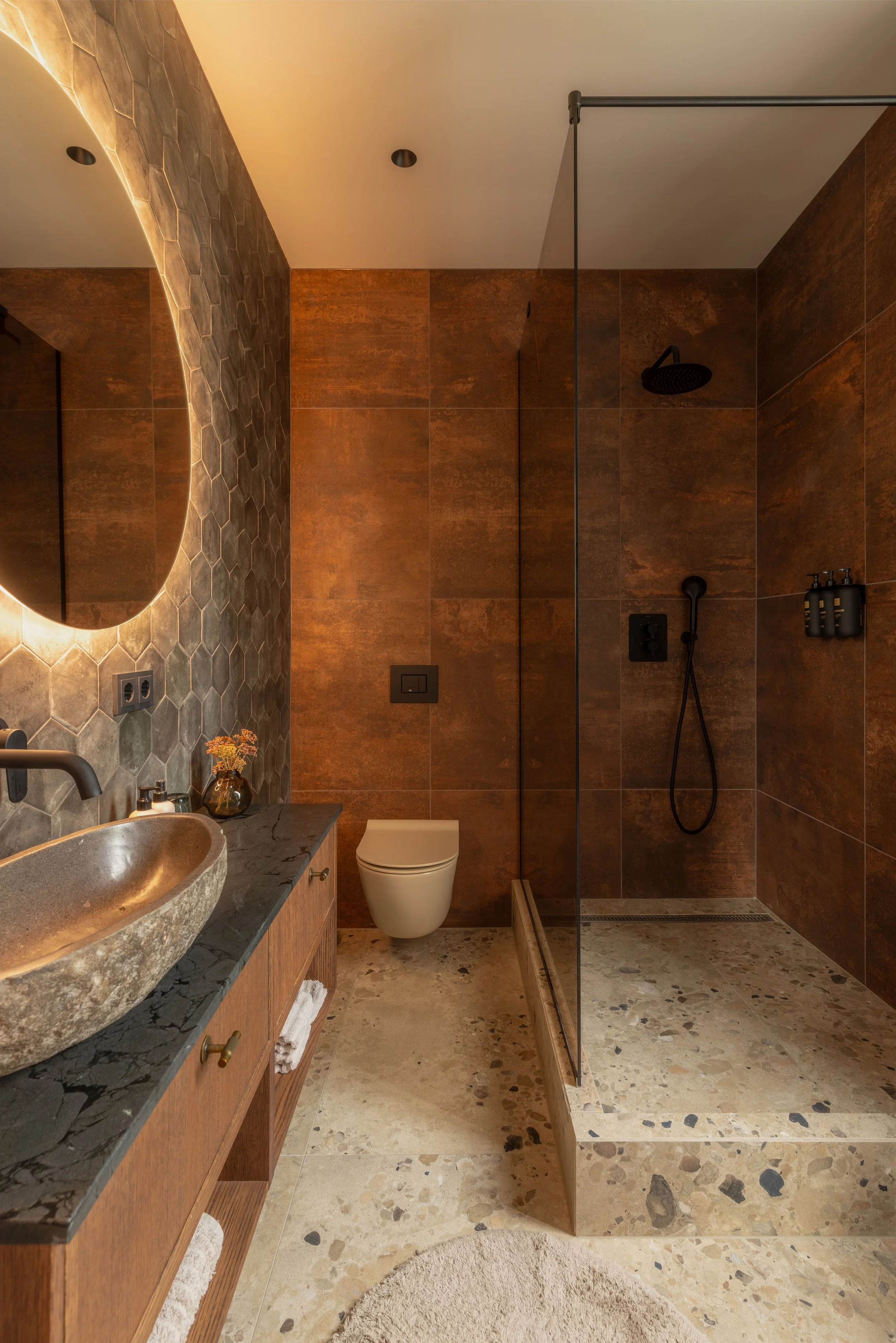 A modern bathroom with a textured stone vessel sink on a dark black marble countertop, wooden vanity with brass handles, a circular beige rug, a wall-mounted toilet, dark brown rust-colored tiles, a glass-enclosed walk-in shower with a black rain sho