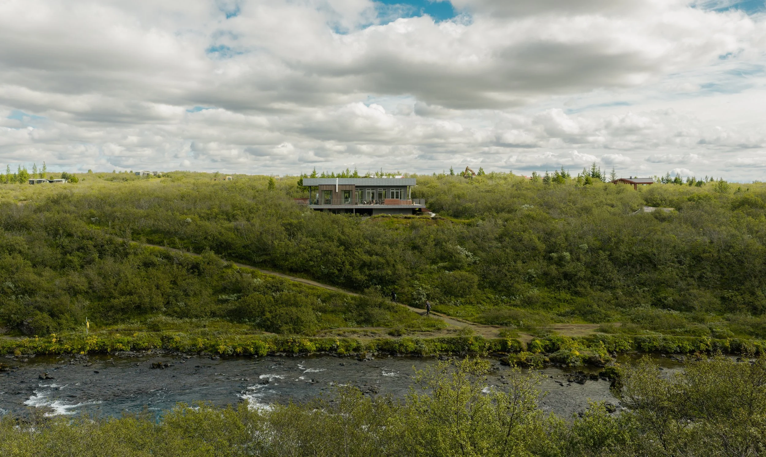A modern house on a hillside surrounded by lush green vegetation, with a river running in the foreground and a partly cloudy sky overhead.