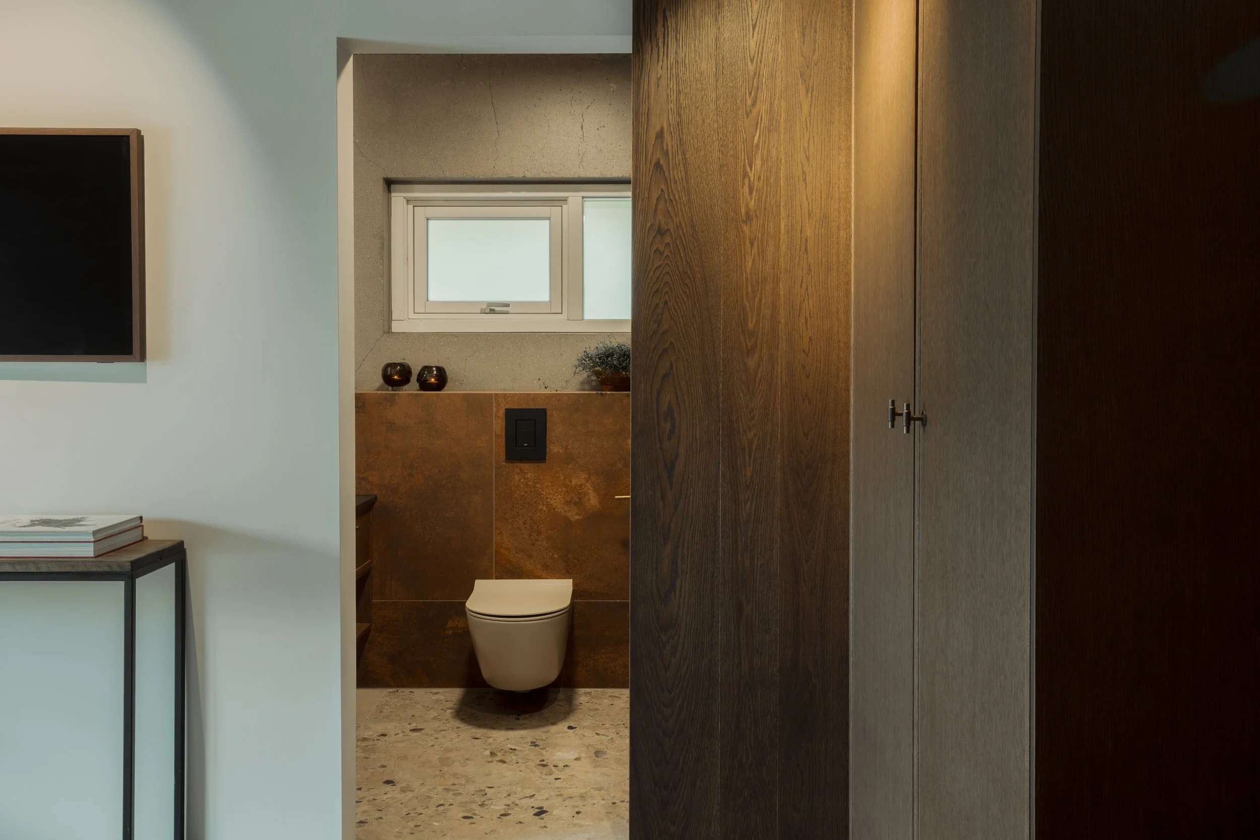 View of a modern bathroom with a white toilet, brown tiled wall, small window, and decorative objects on a shelf.