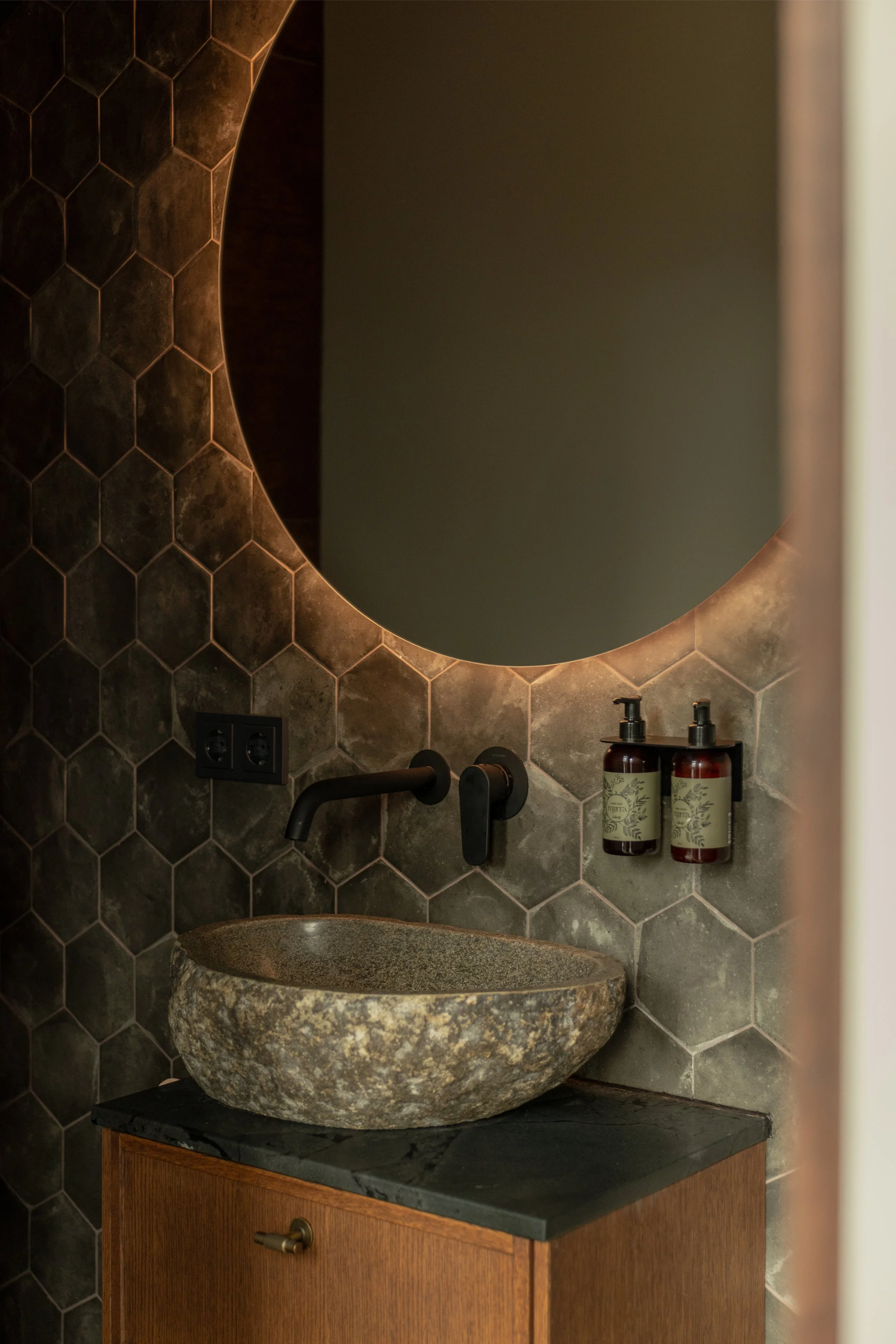 A bathroom with a stone vessel sink on a wooden vanity, hexagonal dark tile wall, black faucet, and two amber soap dispensers mounted on the wall.