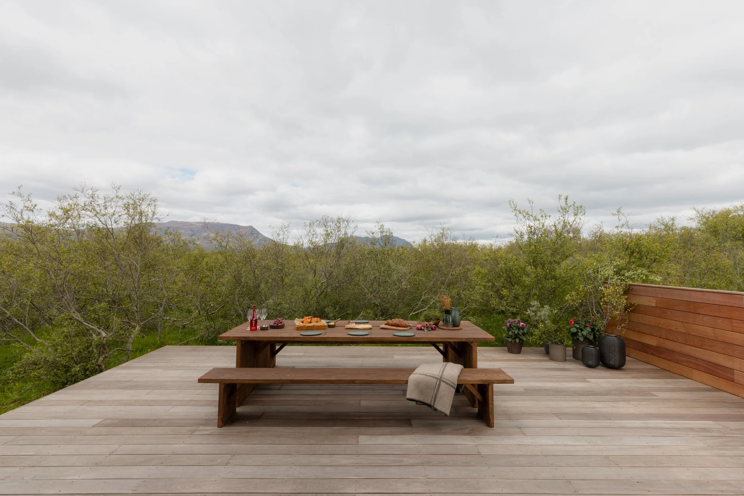 A backyard wooden deck with a dining table set for a meal, surrounded by potted plants and lush green trees, with mountains and a cloudy sky in the background.