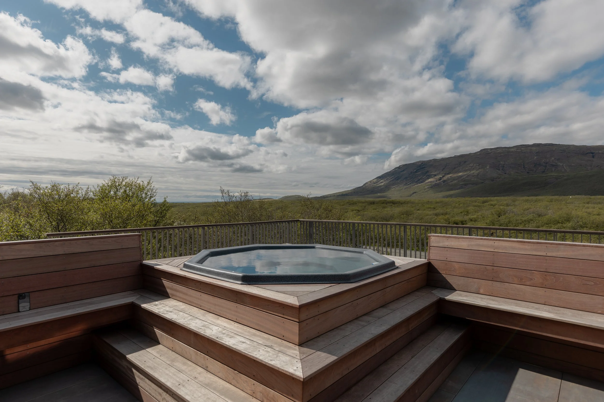 A hot tub on a wooden deck overlooking green hills and a cloudy sky.