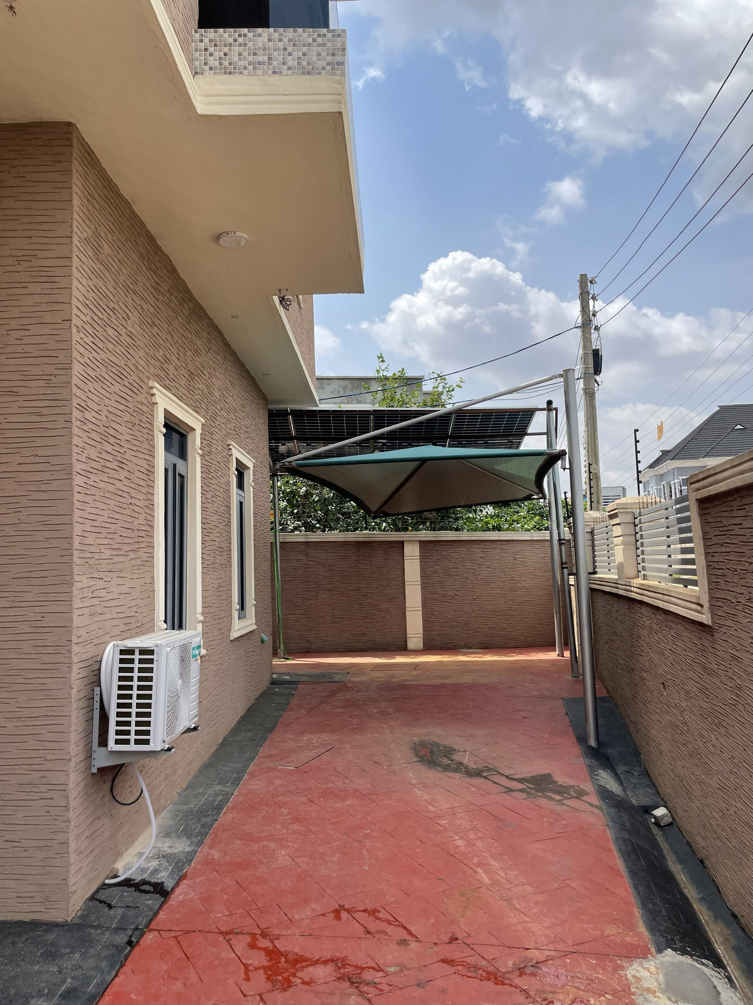 A backyard patio area with a beige house on the left, featuring three narrow windows and an air conditioning unit mounted on the wall. The ground is a reddish stamped concrete with some cracks and wet spots. A black border runs along the base of the house and a black and gray paved walkway is visible on the left side. There's a white fence on the right and a tall utility pole with power lines in the background. Above, the sky is partly cloudy with blue sky and white clouds.