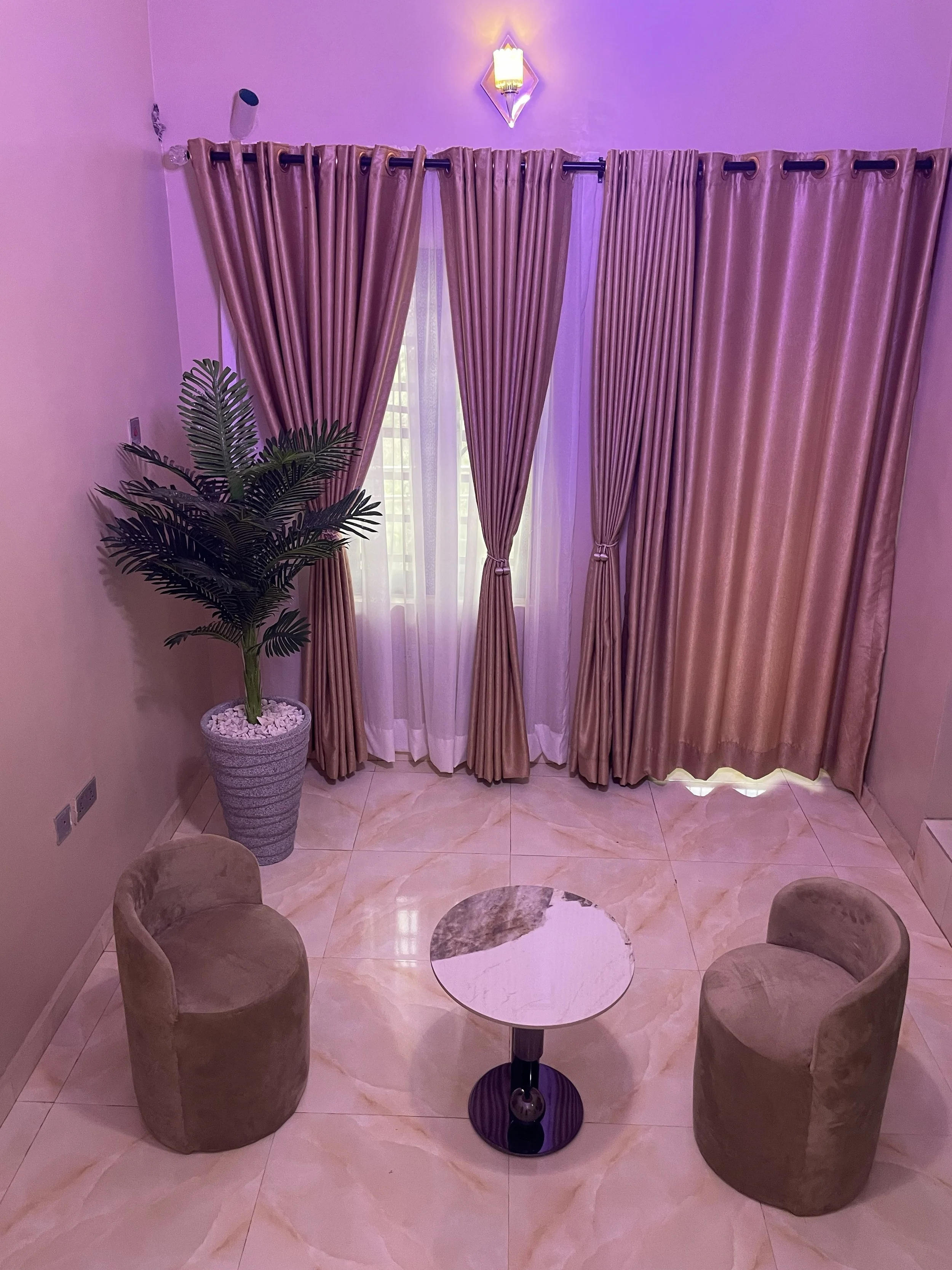 A small round marble table with a black base positioned between three beige plush chairs in a living room. Behind the chairs, there is a window with gold-colored curtains tied back, and purple drapes. To the left of the window, a tall potted plant with dark green leaves sits in a white planter filled with white pebbles. The floor is made of light-colored marble tiles.