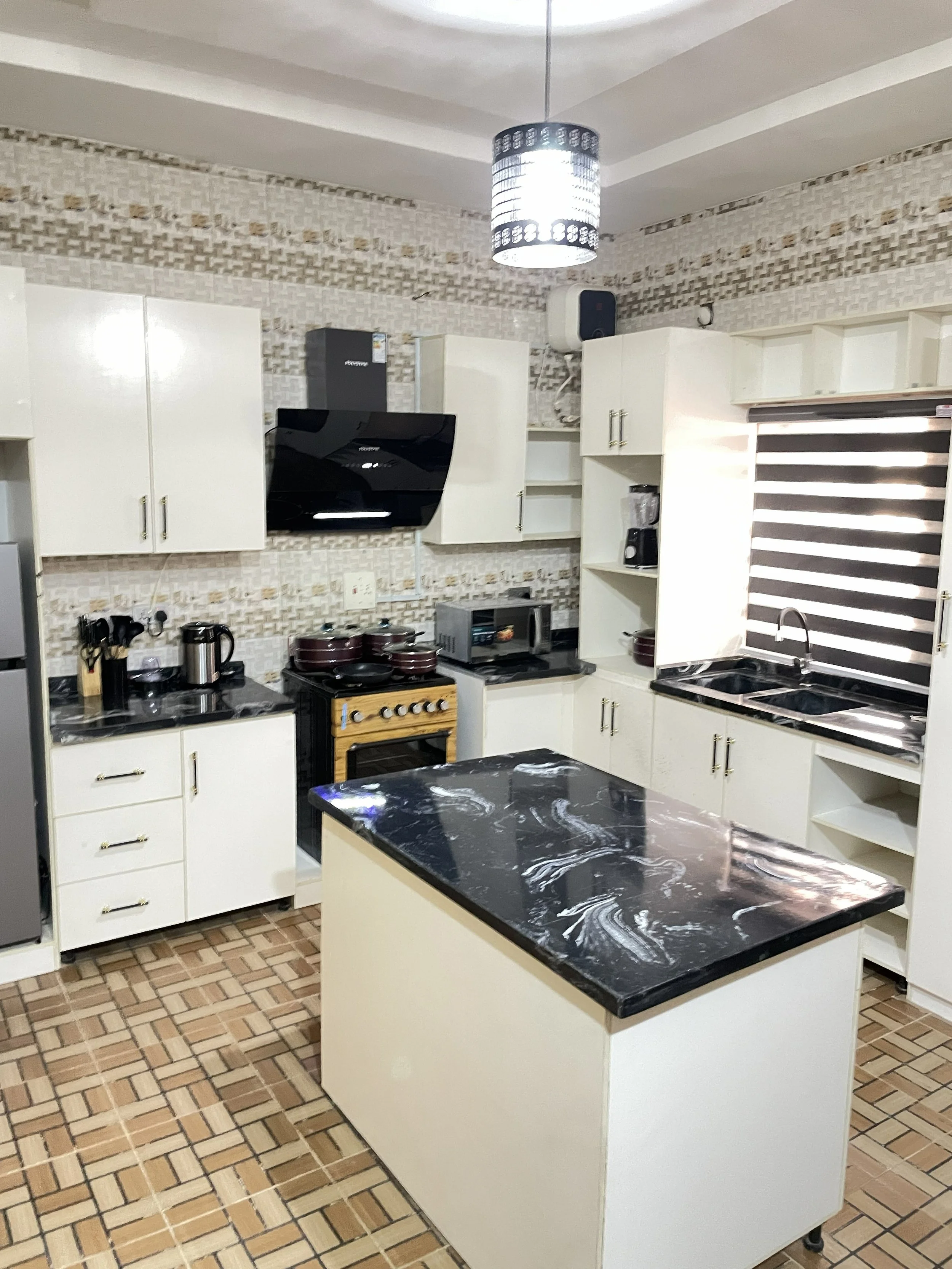 A modern kitchen with white cabinets, black marble countertops, and a central kitchen island. There is a stove, microwave, blender, and other kitchen appliances. The kitchen features tiled walls and a checkered tile floor. A contemporary ceiling light fixture hangs from the ceiling.