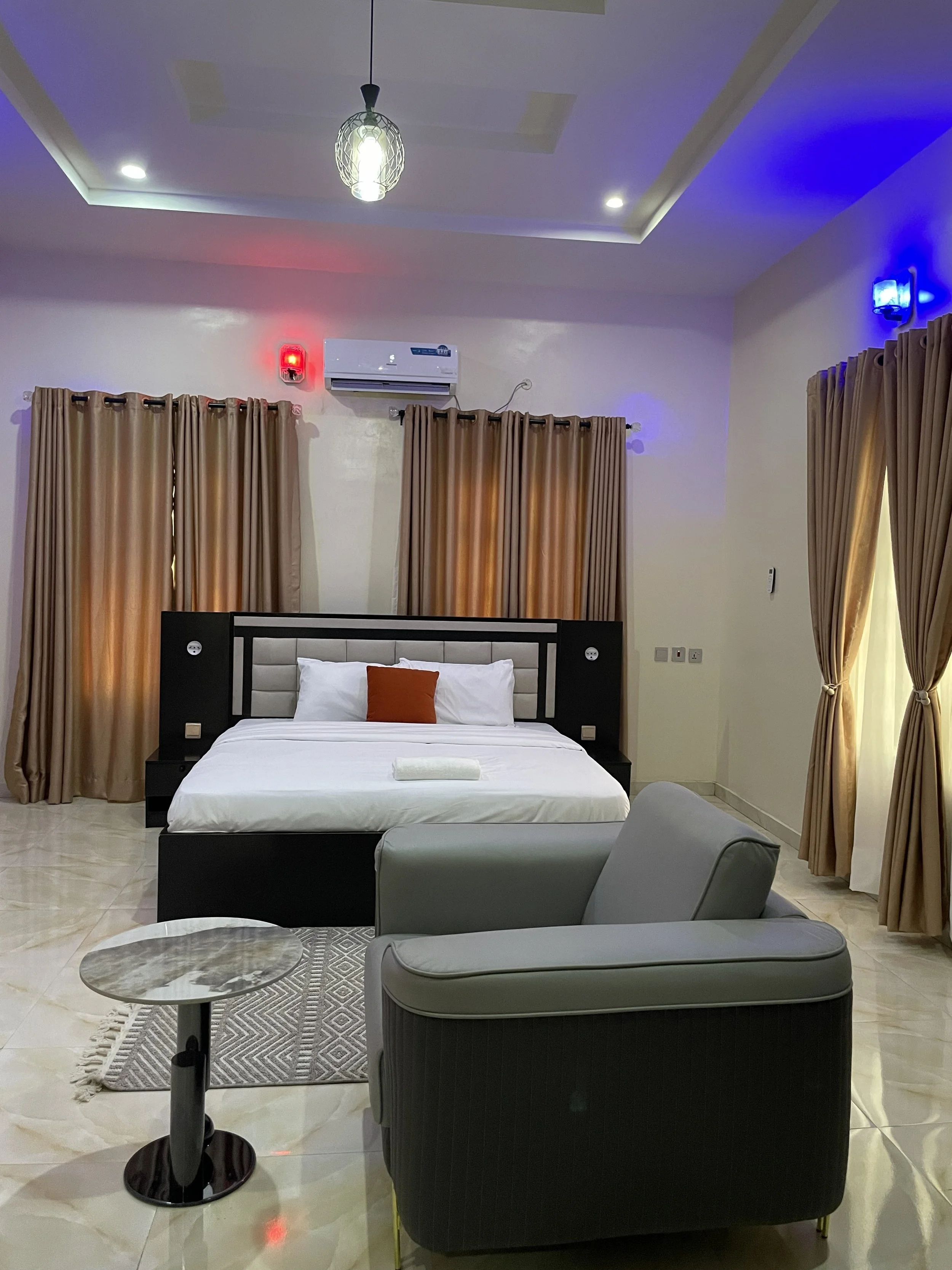 A bedroom with a bed, dark headboard, and pillows, beige curtains, a sitting area with a gray armchair, side table, and patterned rug, air conditioning, and lighting with blue and red accent lights.