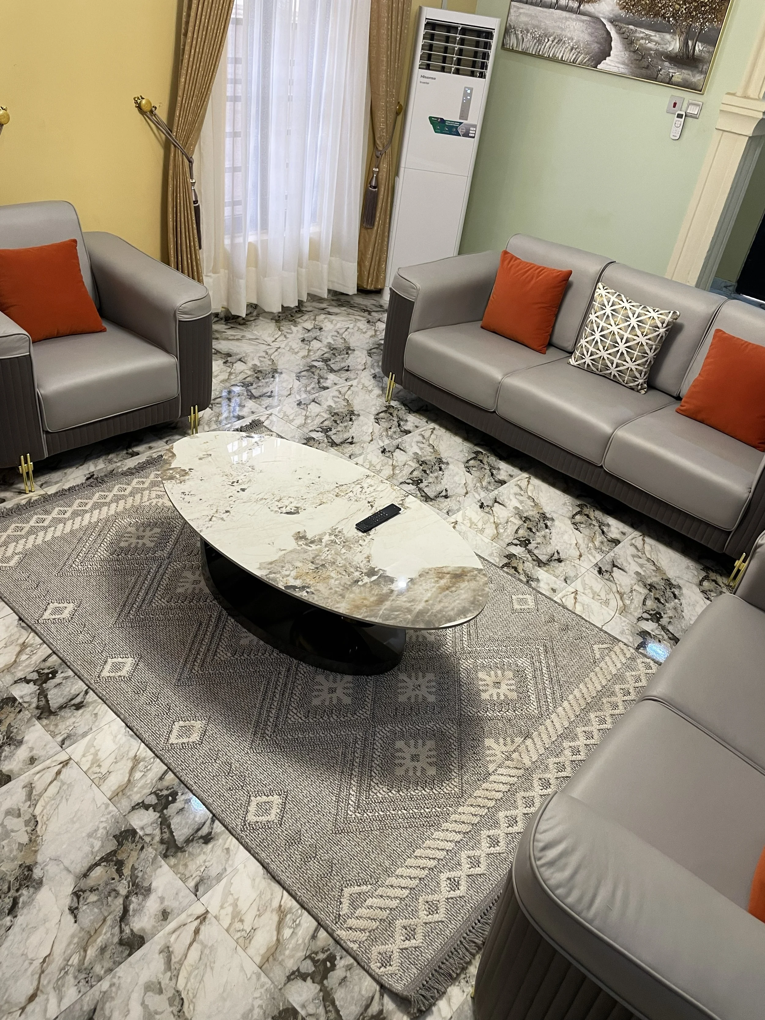 Living room with gray sofas, orange and patterned throw pillows, marble tile floor, marble coffee table, area rug, air conditioner, wall art, curtains, and a remote control on the coffee table.