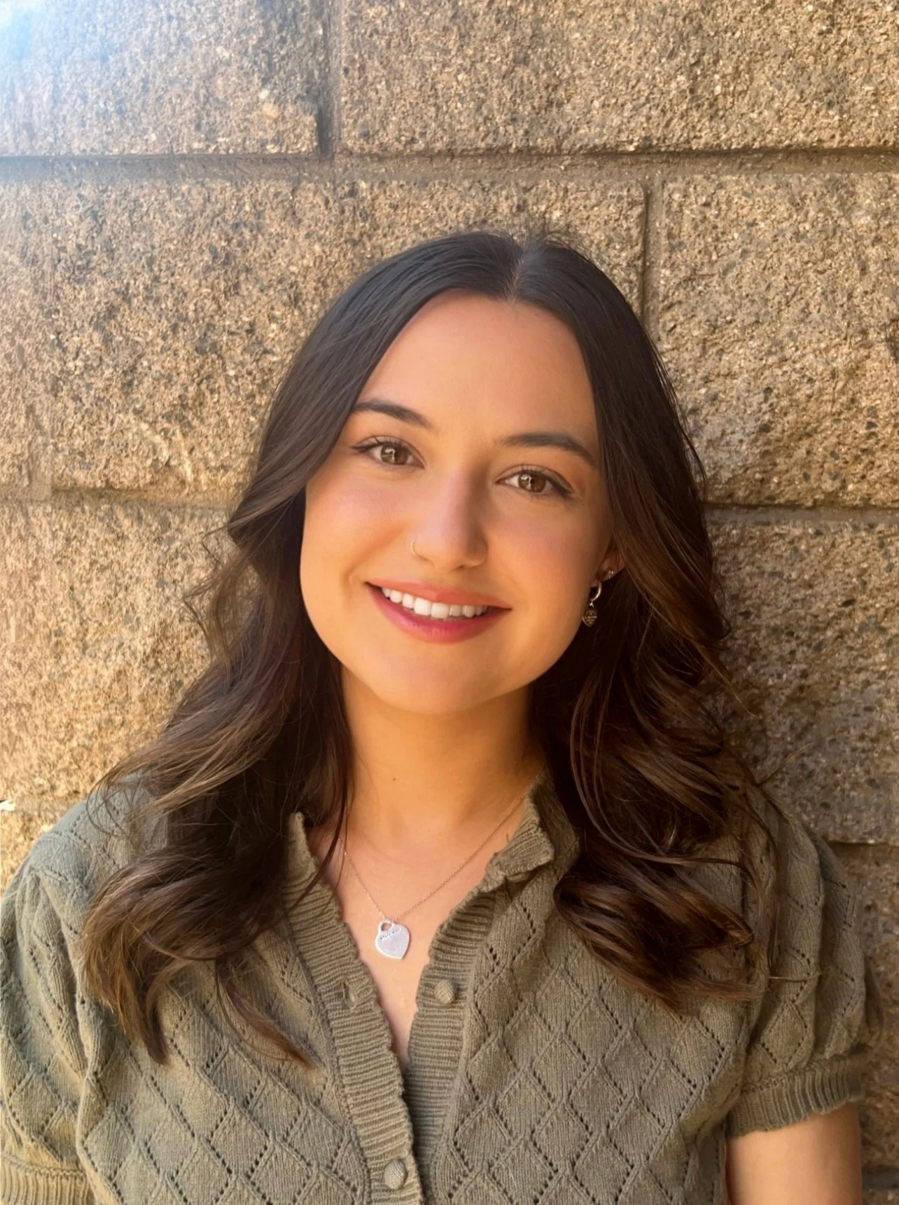A young woman with long, wavy brown hair and brown eyes smiling, standing in front of a tan brick wall. She is wearing a green button-up sweater, a silver necklace with a heart-shaped pendant, and small earrings.