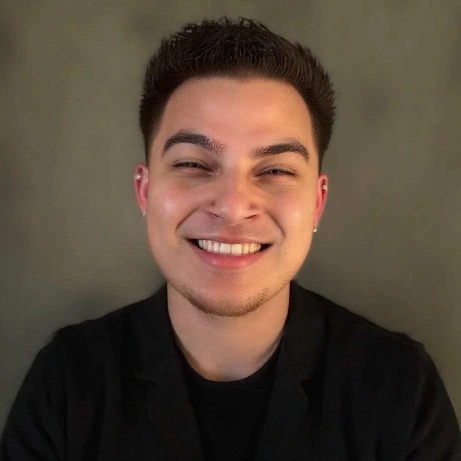 A young man with short dark hair and earrings, smiling broadly, wearing a black top, standing against a plain gray background.