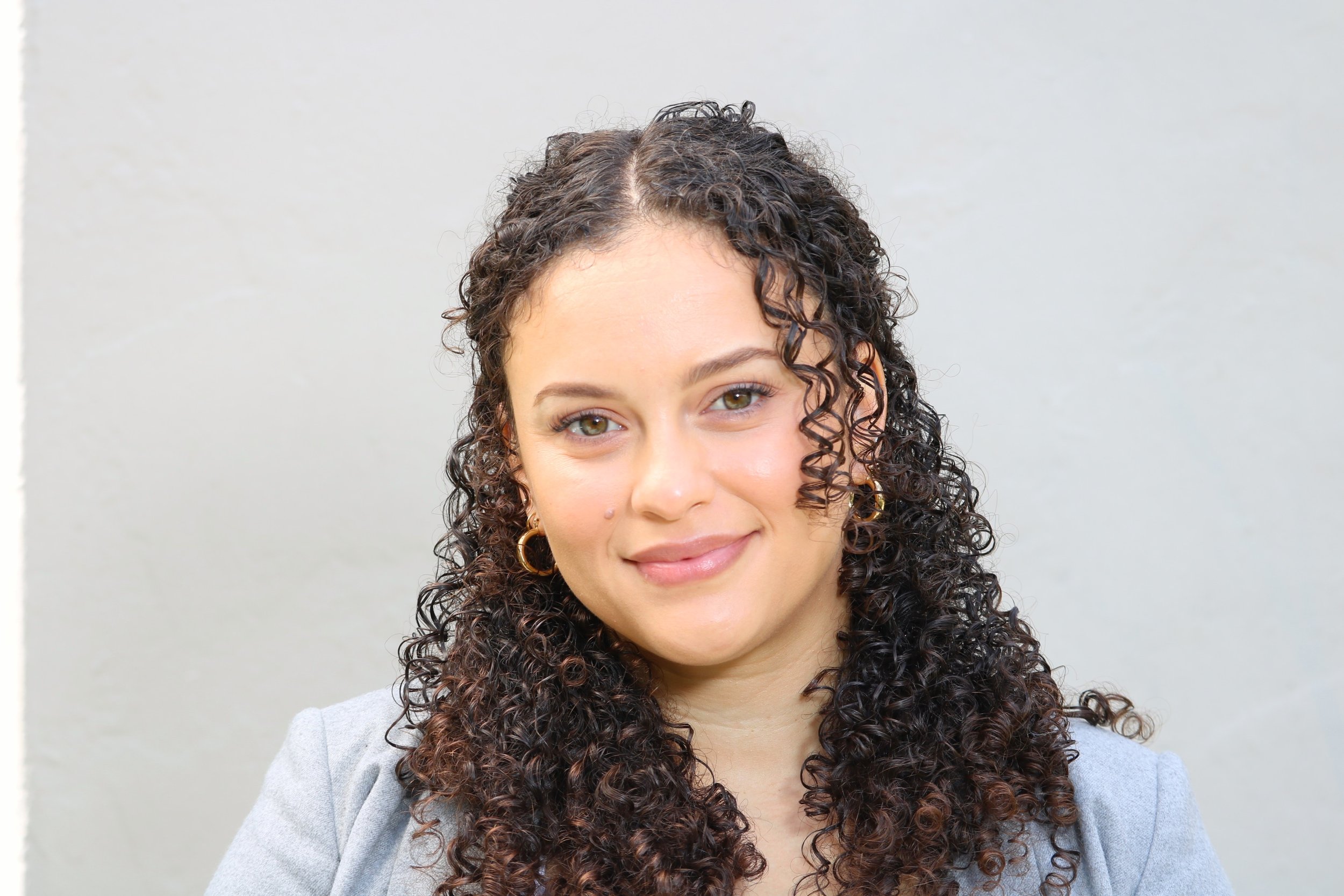 Close-up portrait of a woman with curly dark hair and light skin, smiling, against a plain light background.