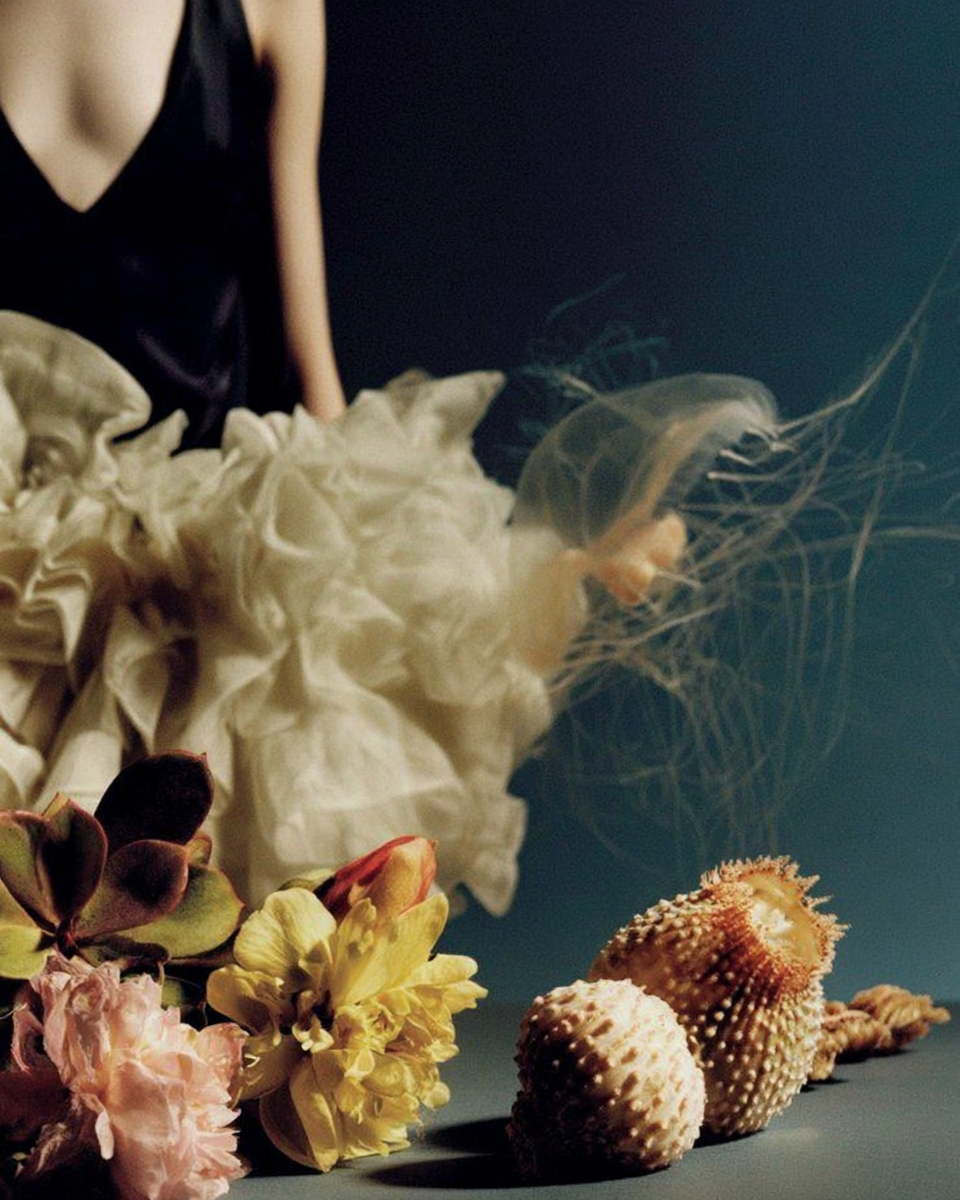 A still life composition featuring flowers, shells, and a silk fabric with a mannequin in the background, against a dark background.