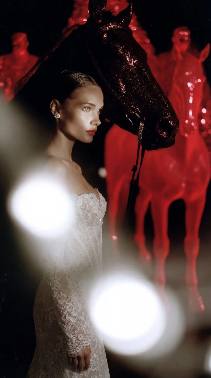 A woman in a white lace dress standing next to a black horse with sparkly features, with red sculptures of horses in the background at night.