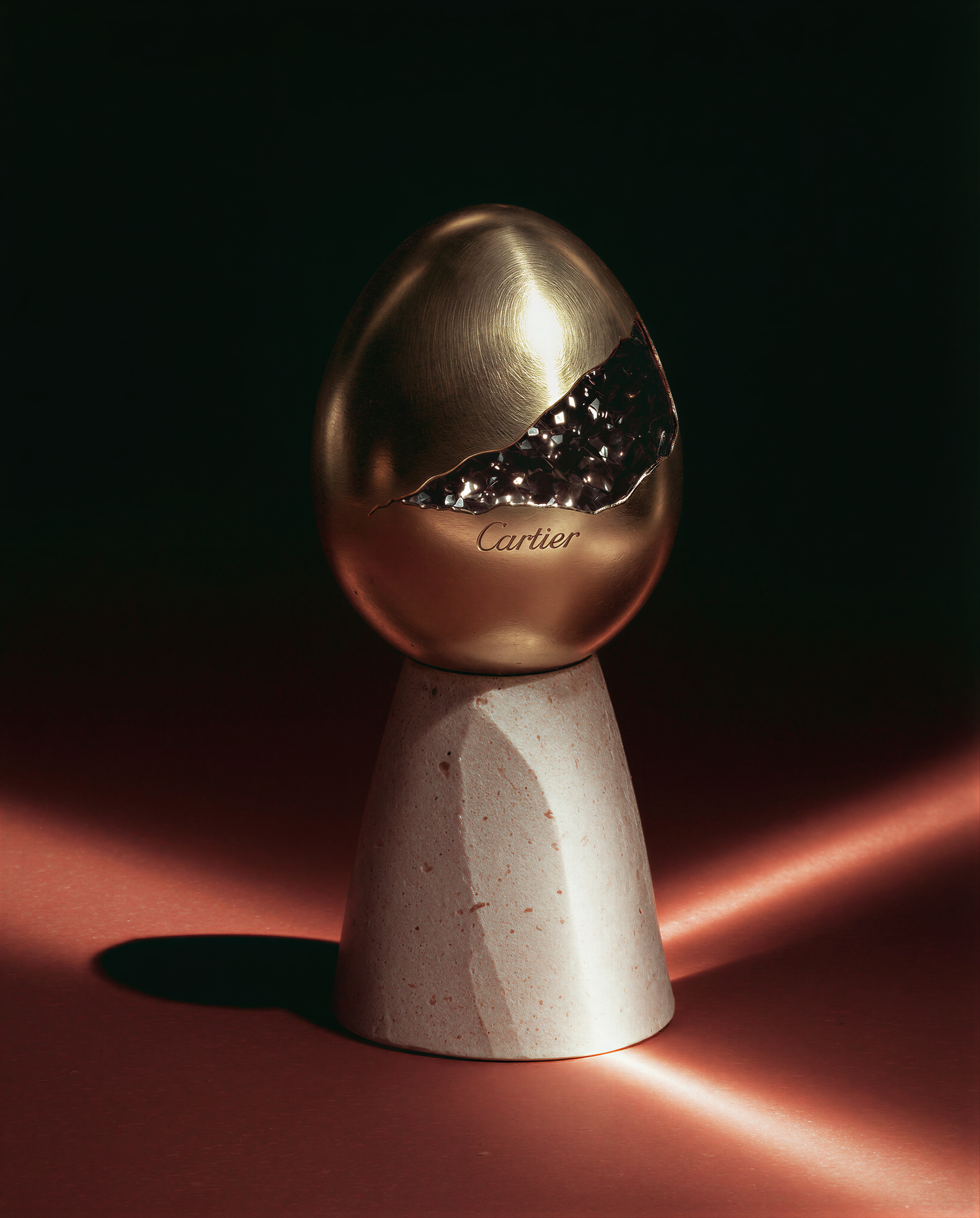 A luxurious golden egg sculpture with a crack revealing dark interior and the word 'Cartier' inscribed, placed on a stone pedestal, with soft lighting and shadows.