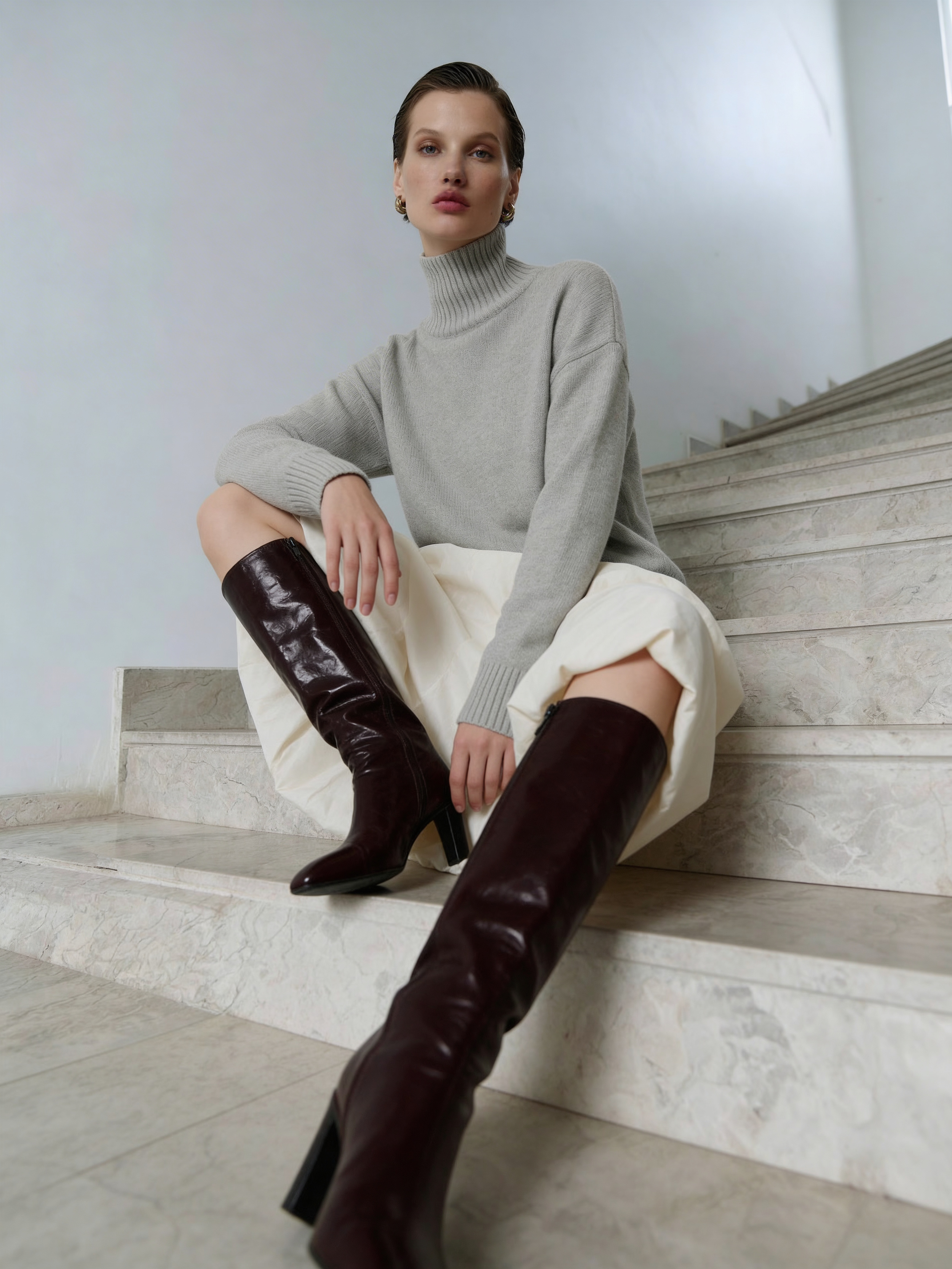A woman sitting on marble stairs wearing a gray turtleneck sweater, cream-colored skirt, and knee-high black leather boots.