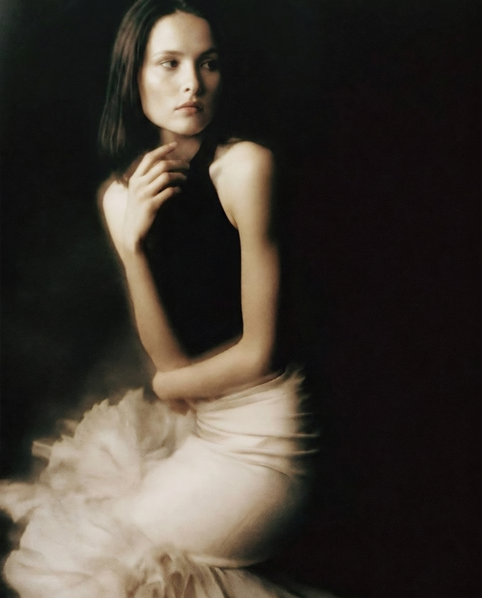 A woman with shoulder-length dark hair and fair skin, wearing a sleeveless black top and white skirt with ruffles, posing against a dark background, looking to the side with her hand near her chin.