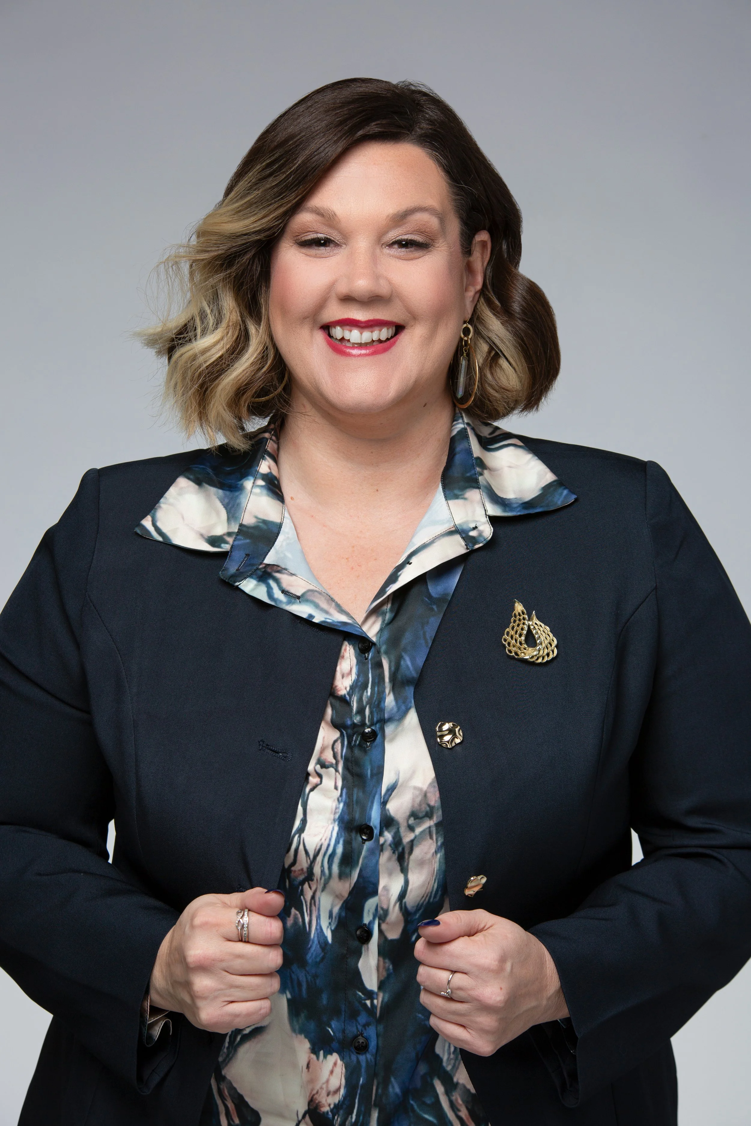 A woman smiling, wearing a dark blazer with decorative pins and a blouse with a watercolor pattern, posed in front of a light gray background.