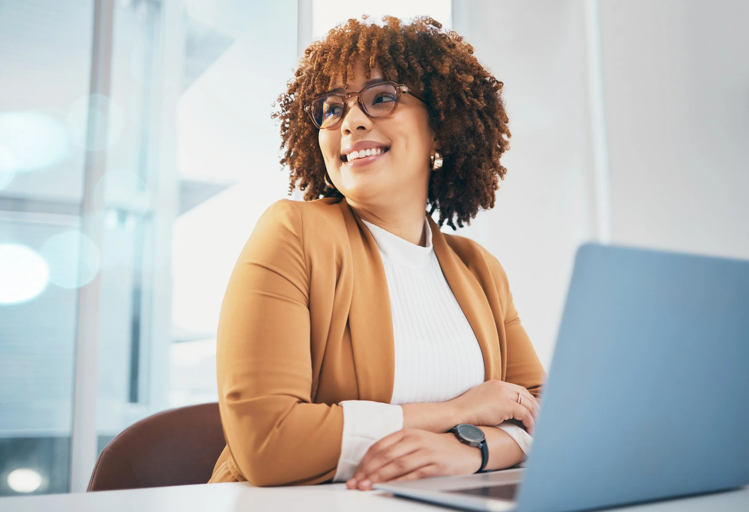 A smiling woman with curly hair, glasses, and earrings, sitting at a table with a laptop in a bright office setting.