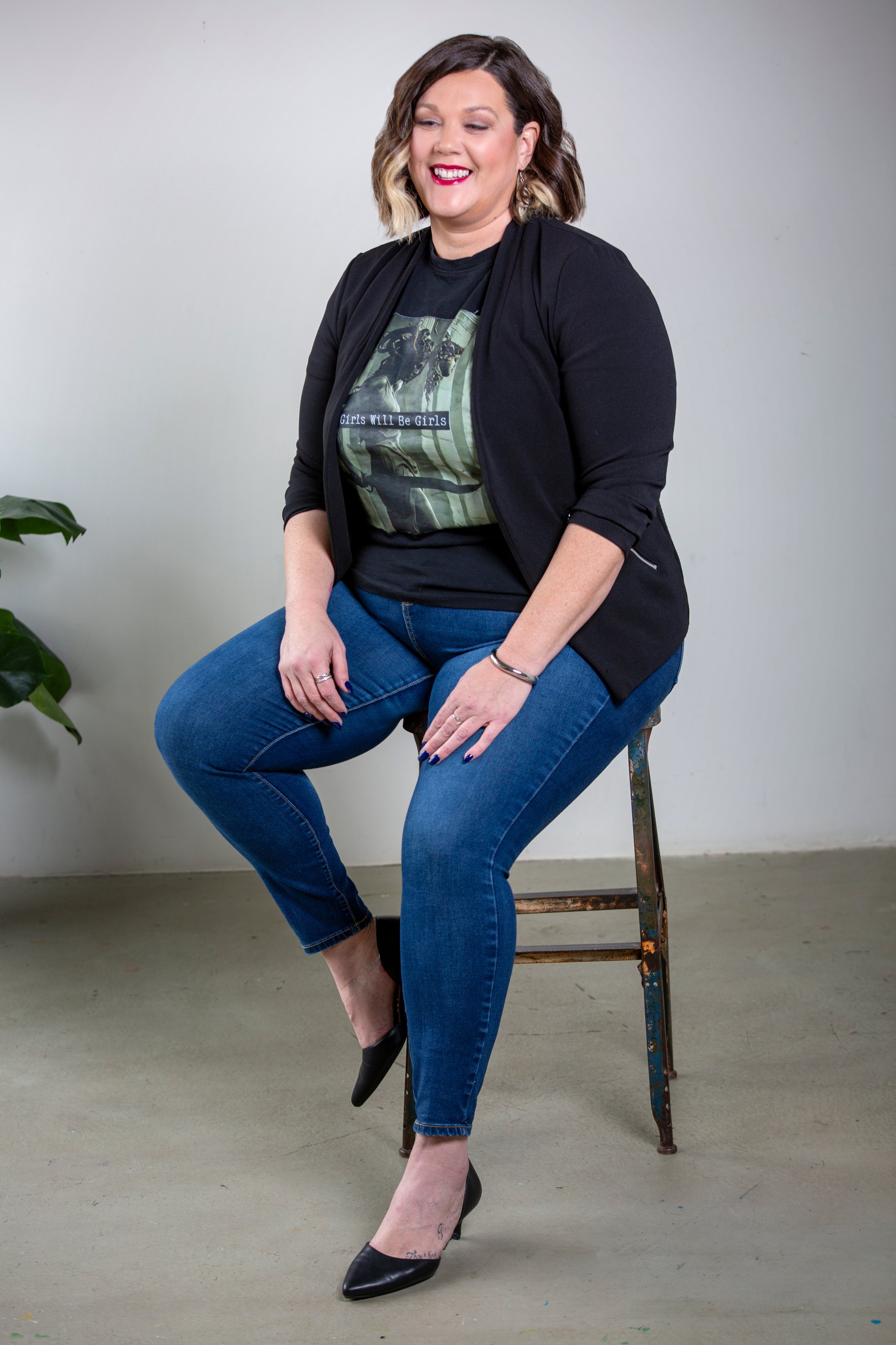 A woman with shoulder-length wavy brown hair and blonde highlights, wearing a black blazer, graphic T-shirt, blue jeans, and black high heels, sitting on a rustic wooden chair against a plain light gray background, smiling with makeup including pink lipstick.