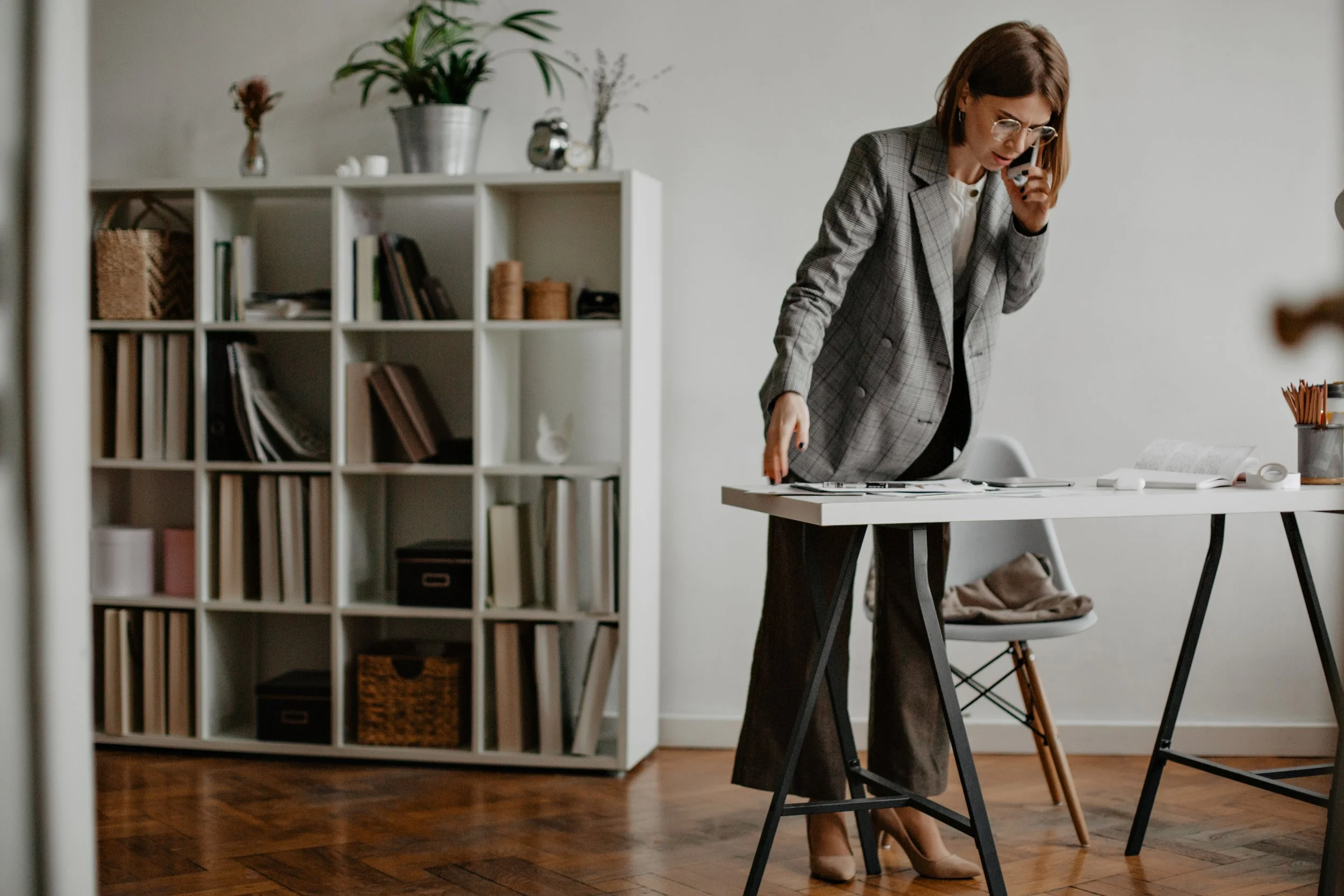 A woman in business attire is standing at a desk, talking on a cellphone, with documents and office supplies on the table, in a modern office setting.