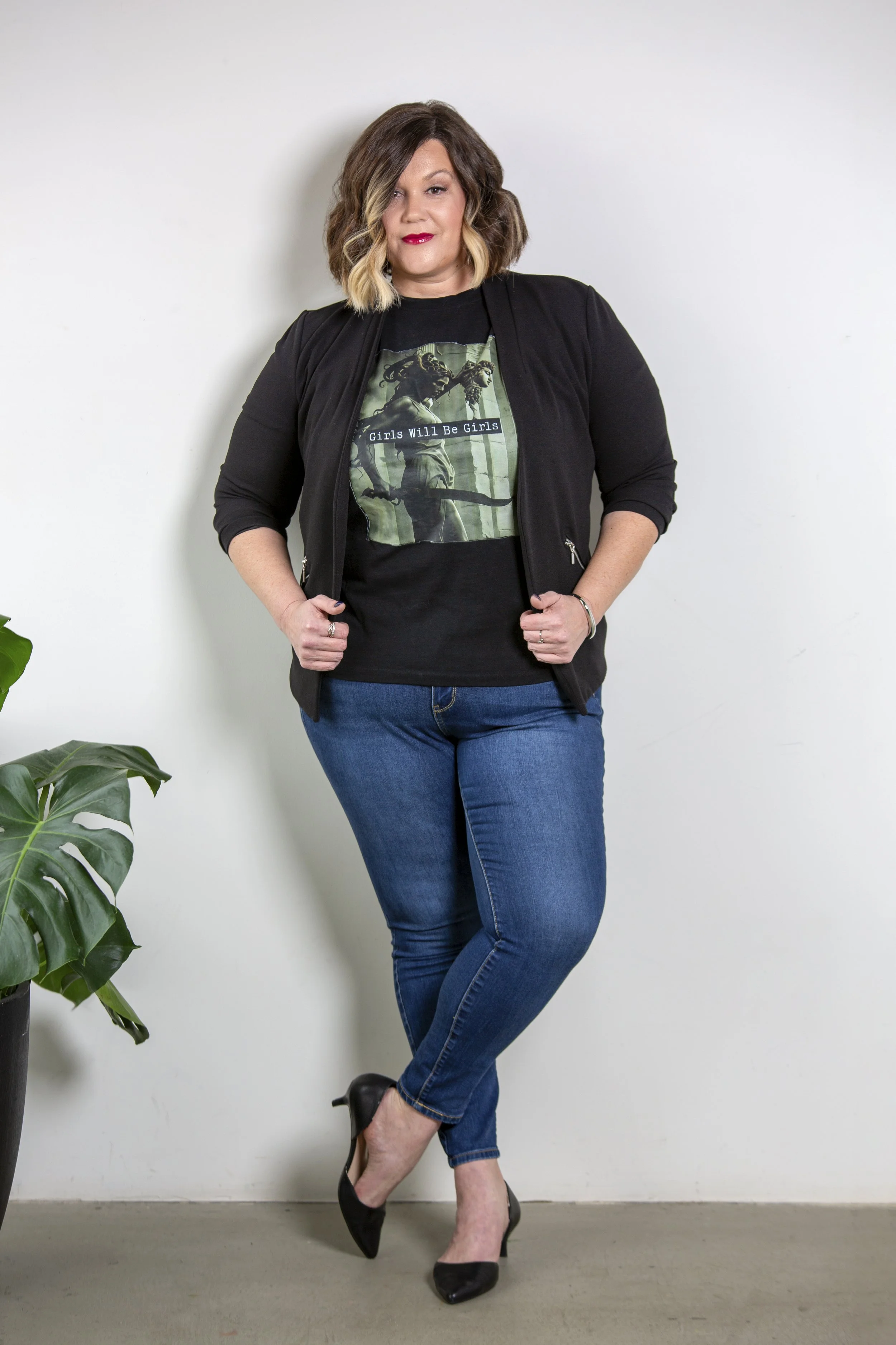 A woman with shoulder-length brown and blonde hair wearing a black blazer, a black graphic t-shirt with the text 'Girls Will Be Girls' and an image of mythical creatures, blue jeans, and black high heels, standing against a white wall with a green plant visible on the left.