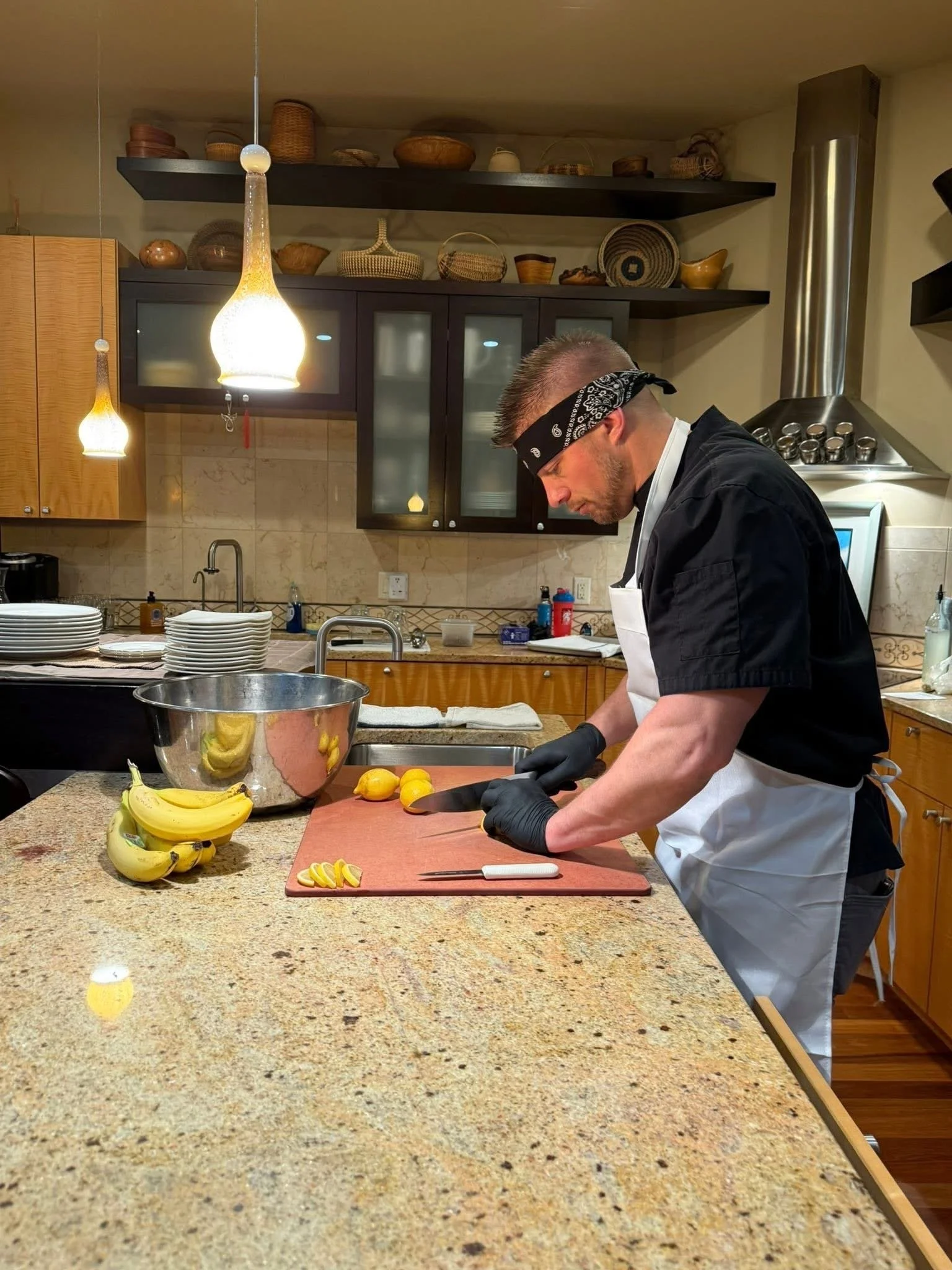 A man wearing a black bandana, black gloves, and a white apron is chopping lemons on a red cutting board in a kitchen with a beige granite countertop. There are yellow bananas on the counter, and kitchen cabinets and open shelves with baskets and dis
