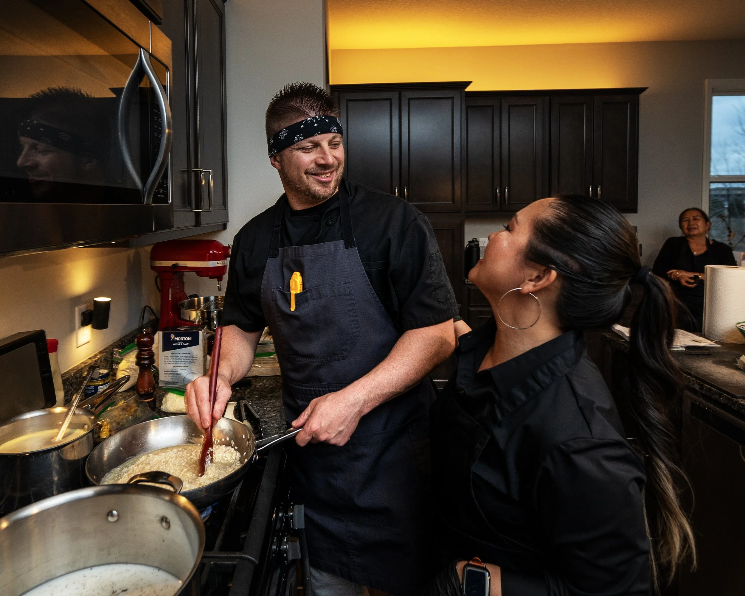 A man and a woman cooking together in a kitchen with dark cabinets. The man is smiling and wearing a black shirt, an apron, and a black bandana, while the woman, with only part of her face visible, is wearing hoop earrings, and her hair is tied back.