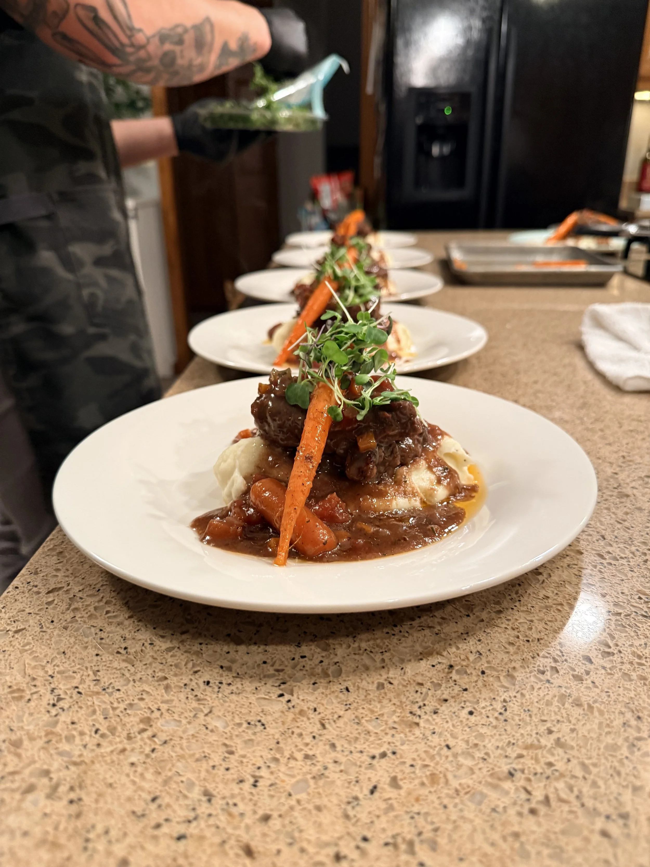 Plated serving of beef and mashed potatoes garnished with microgreens and a carrot, in a restaurant kitchen setting.