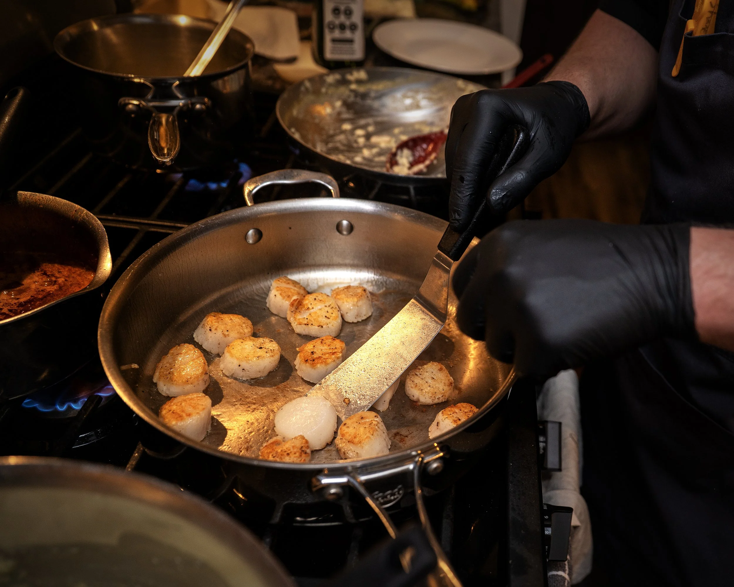 A person wearing black gloves is cooking scallops in a frying pan, with other pots and dishes visible in the kitchen.