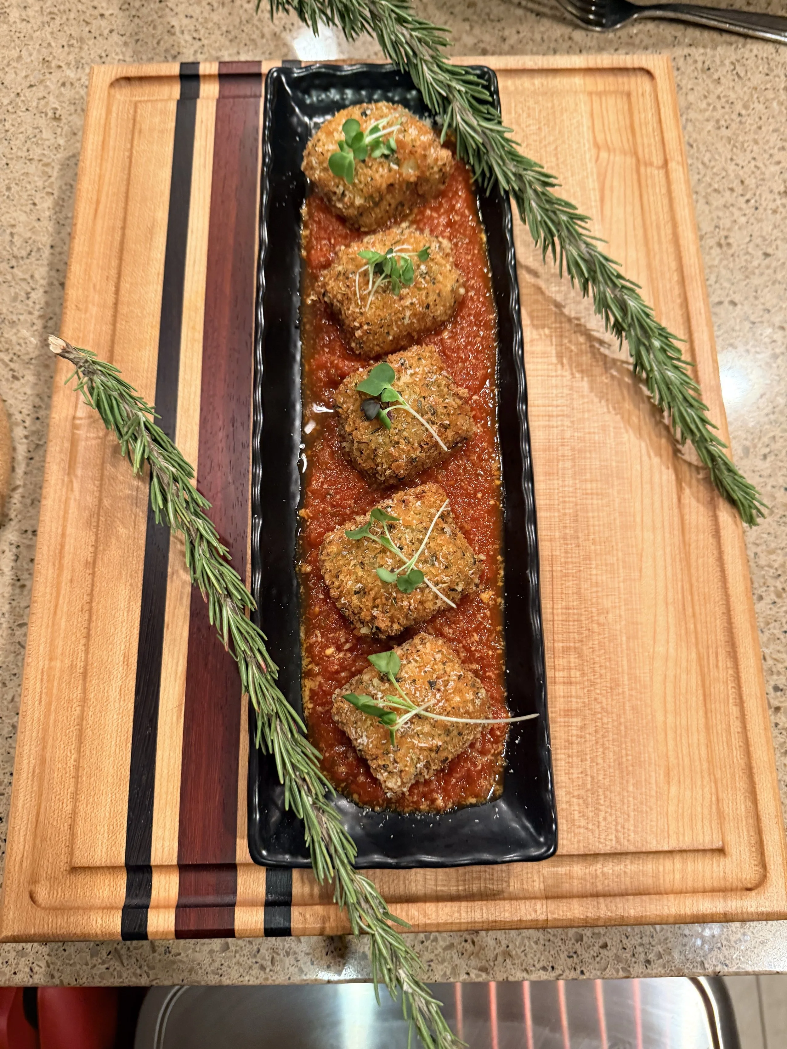 Four croquettes with green microgreens on top, served on a black rectangular plate with tomato sauce, garnished with rosemary sprigs on the side, placed on a wooden cutting board.