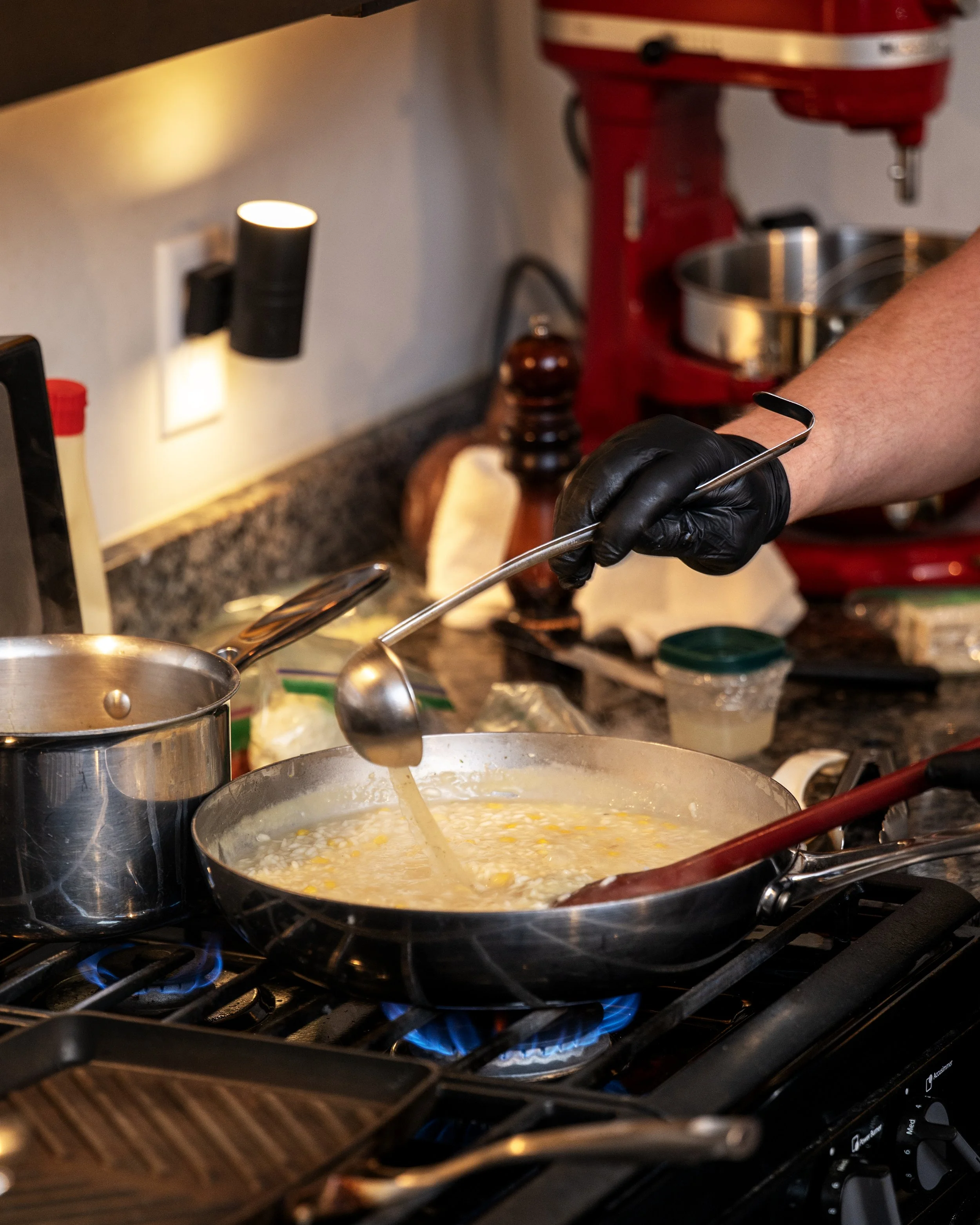 A stove with a boiling pot of creamy soup with corn and other ingredients, and a person wearing a black glove stirring it with a spoon, in a kitchen with various utensils and appliances.