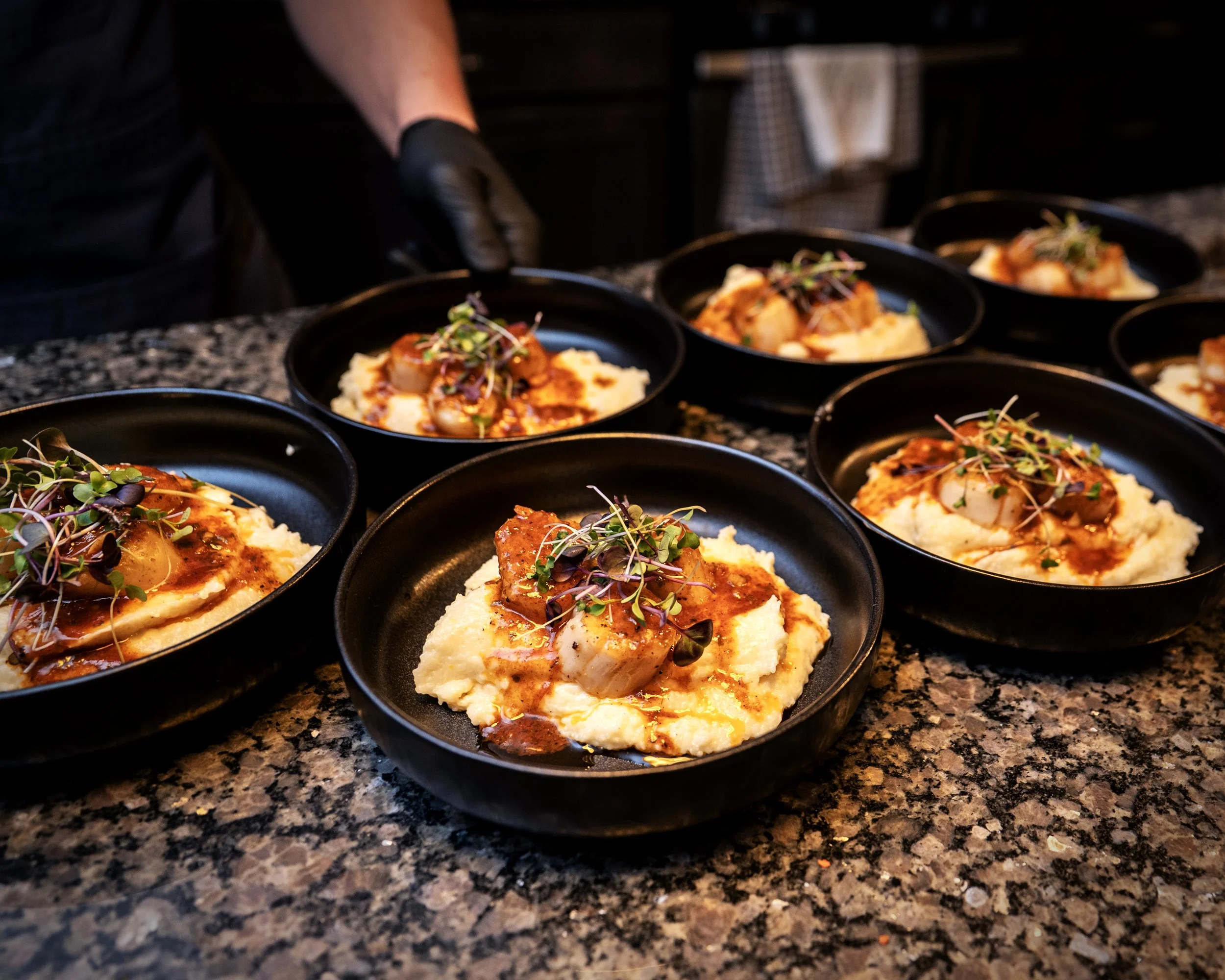Several black bowls filled with mashed potatoes topped with a spicy meat stew and garnished with microgreens, set on a granite countertop at a restaurant kitchen.