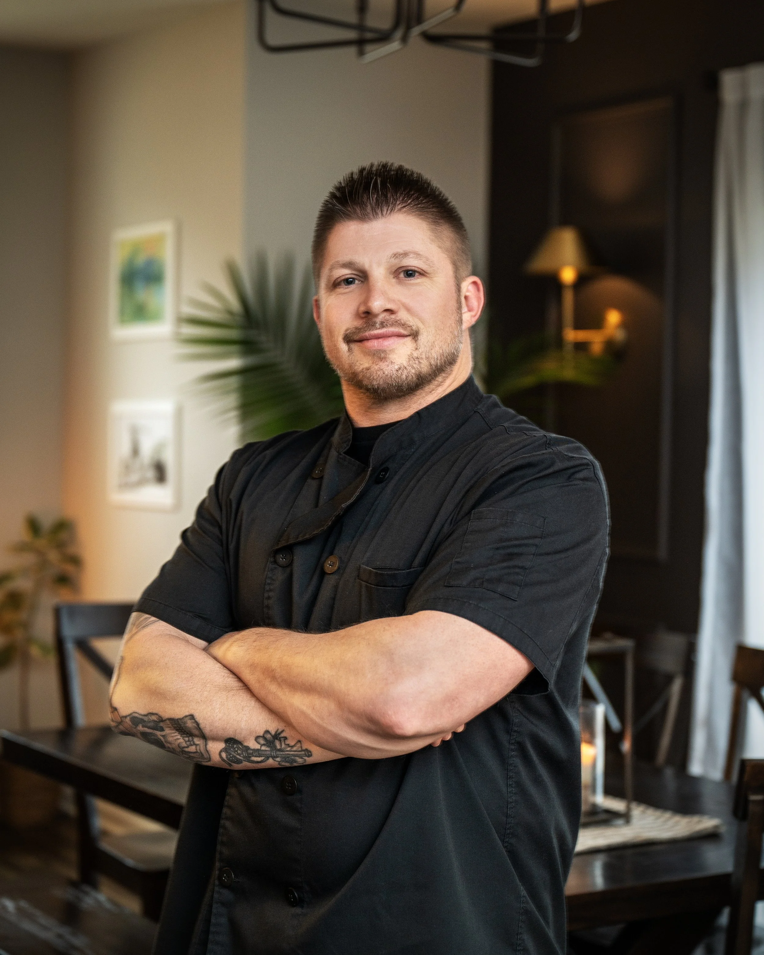 A man with short dark hair, a beard, and tattoos on his forearm, standing with arms crossed, wearing a black button-up shirt, in a cozy indoor setting with warm lighting, a plant, and artwork on the wall behind him.