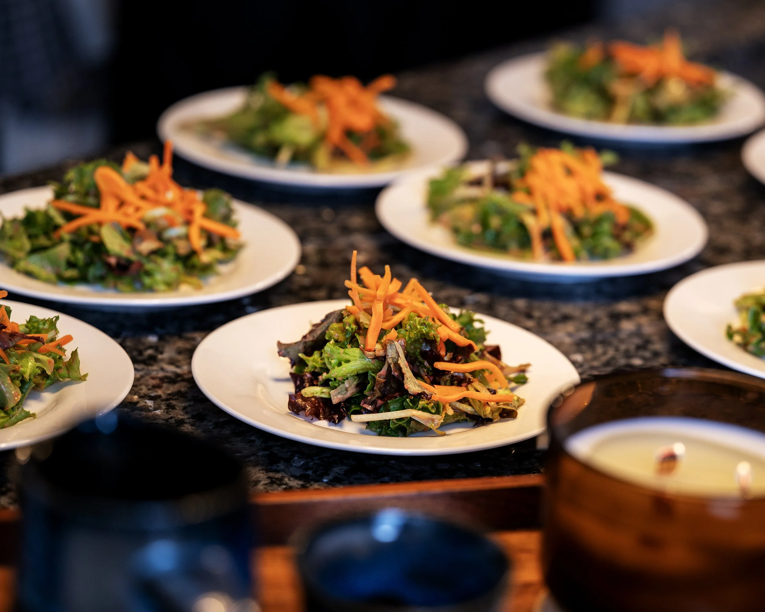 Multiple small white plates filled with salad greens topped with shredded carrots, set on a dark countertop.