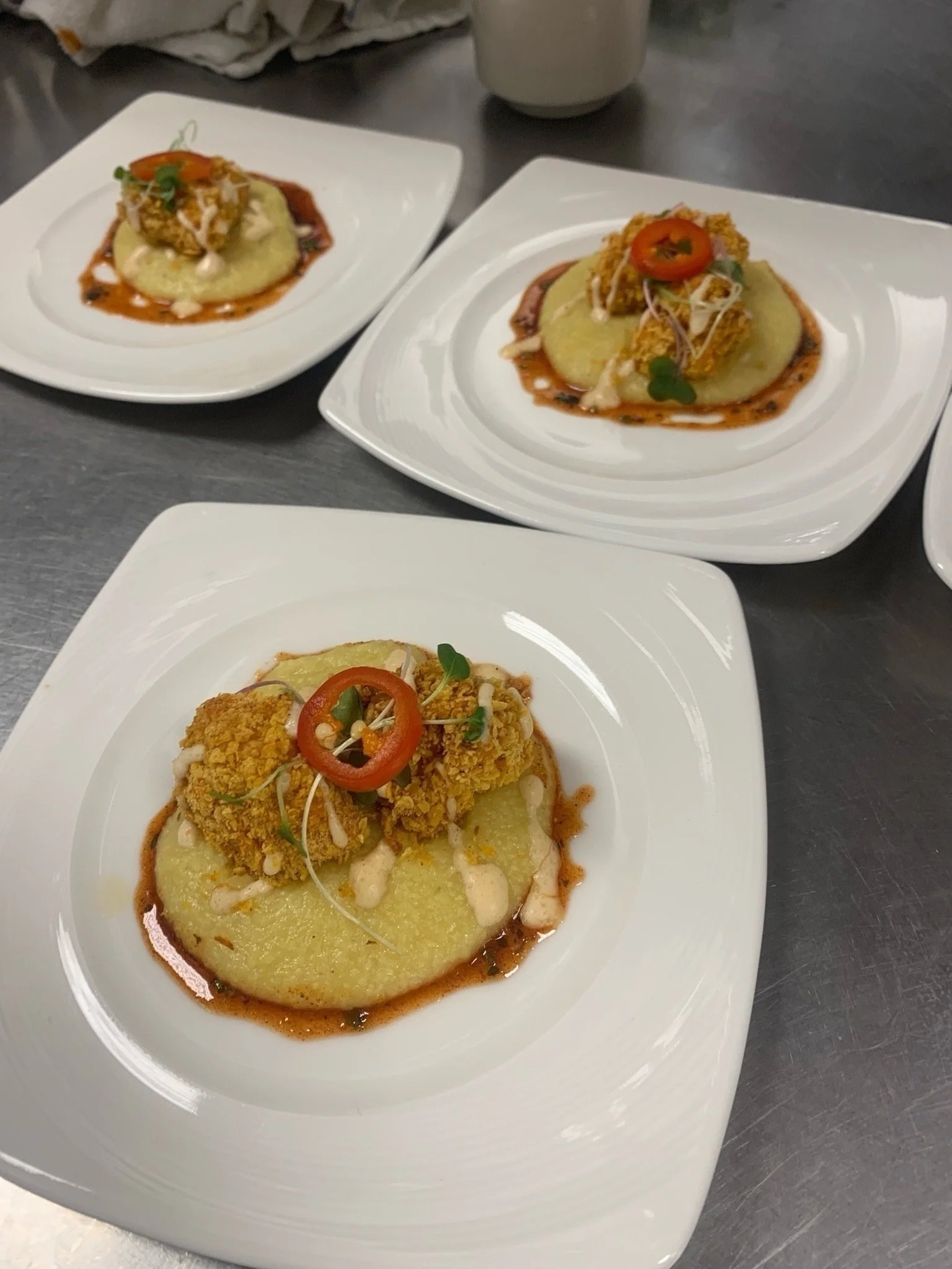 Close-up of plated food featuring a large, round, yellow polenta cake topped with a crispy, breaded chicken fillet, garnished with a red pepper ring, microgreens, and drizzled with sauce on a white square plate.