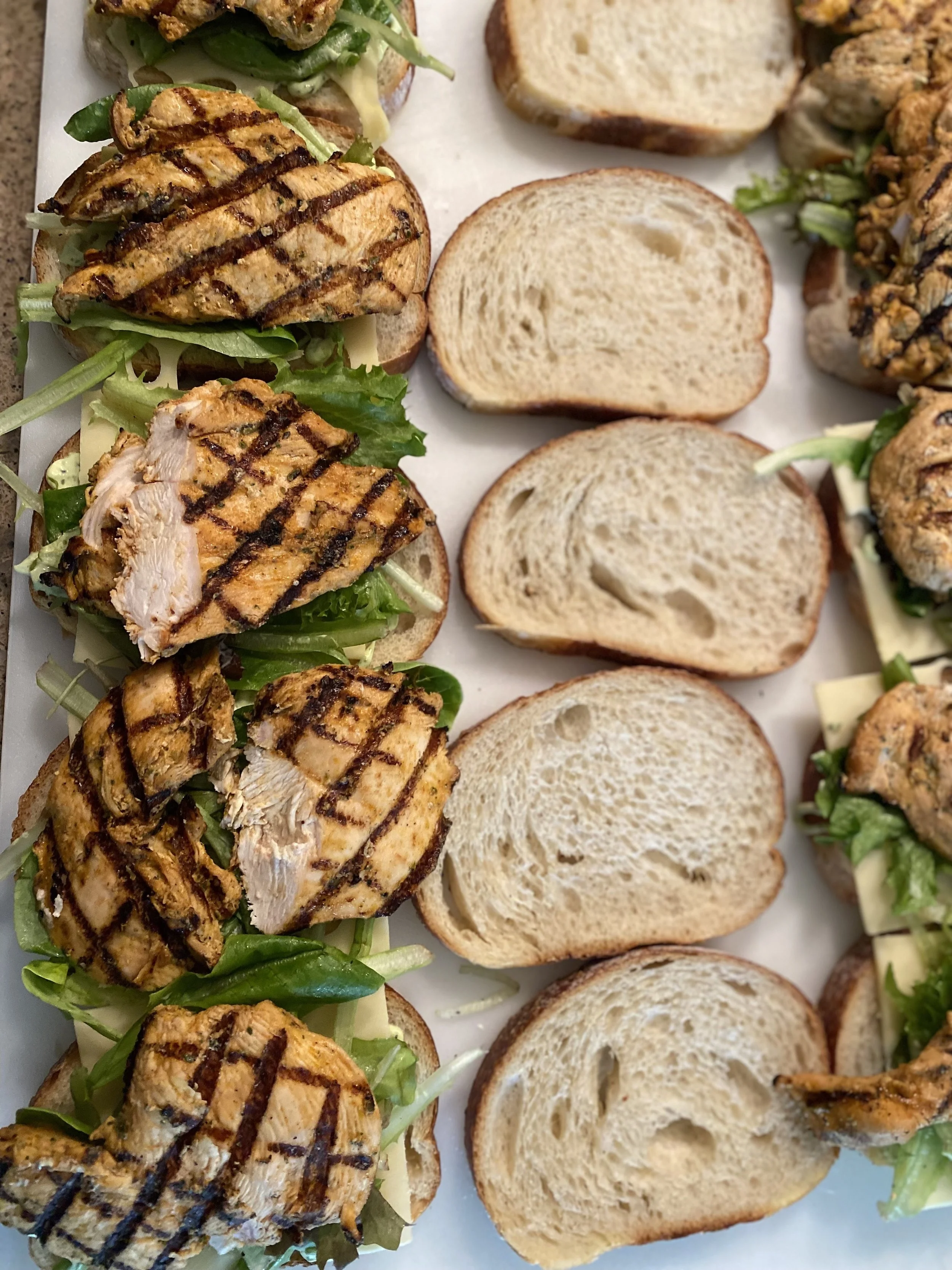 Open-faced chicken sandwiches with grilled chicken breast, lettuce, and sandwich buns on a white platter.