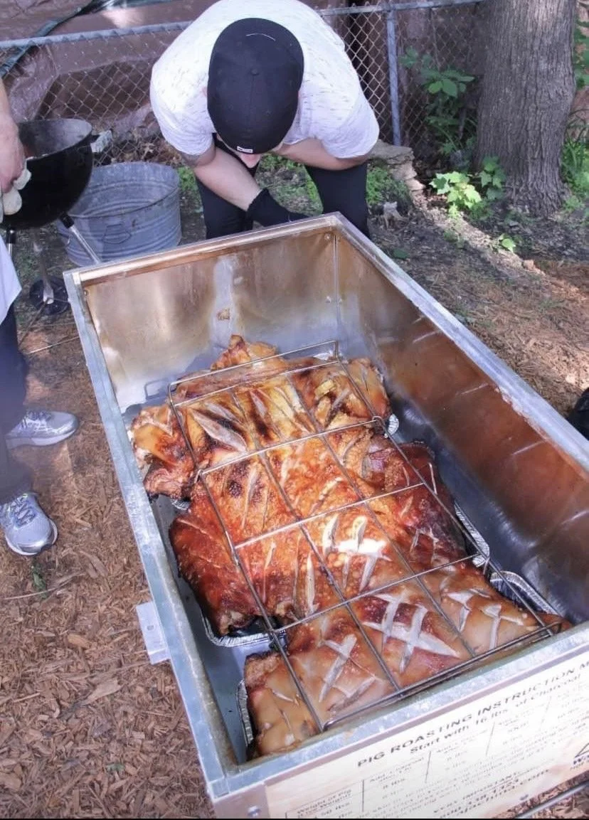 A person tending to a large pig roasting in a metal pit with a grill on top, in an outdoor setting.