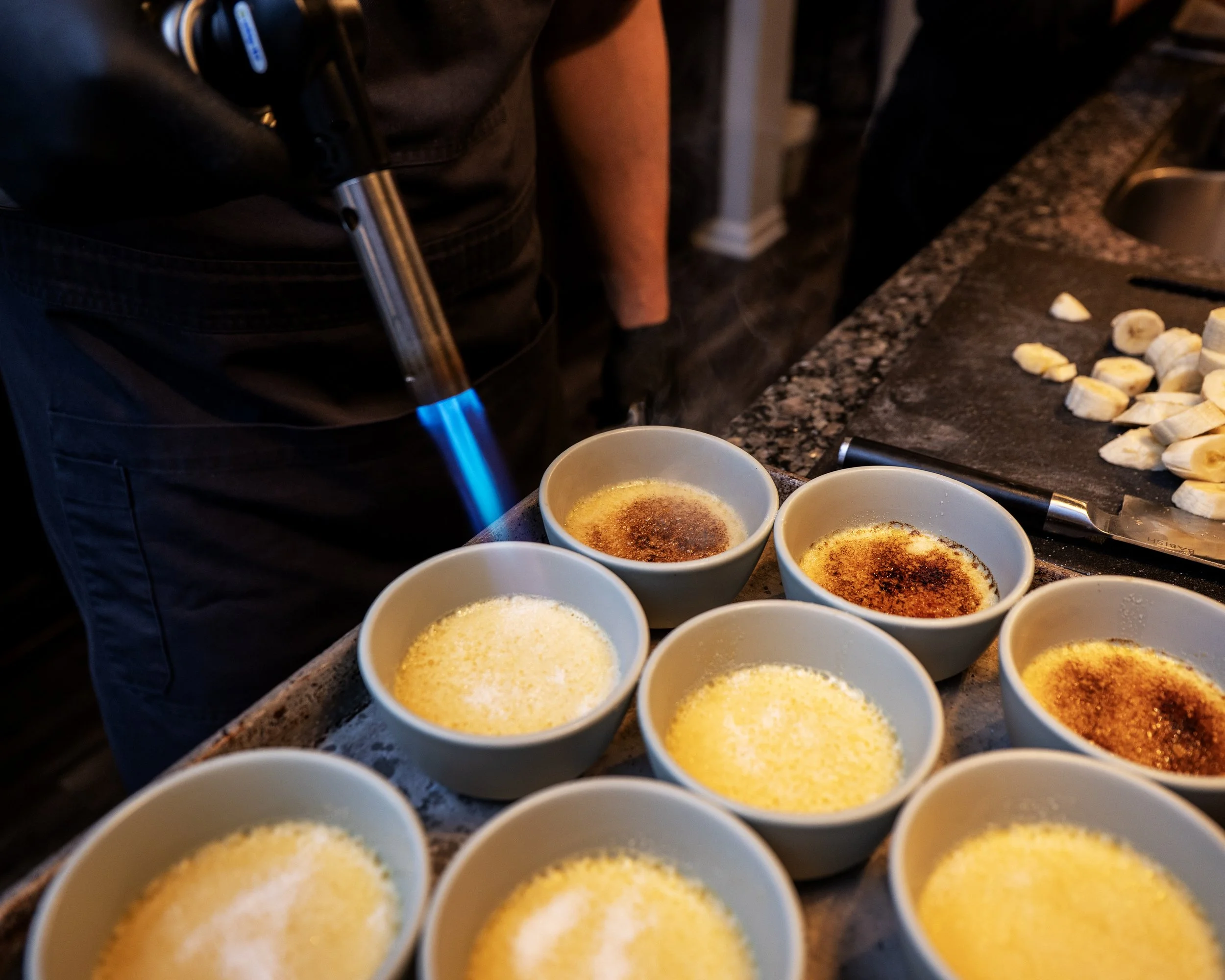 Several cups of crème brûlée with caramelized sugar toppings on a kitchen counter, bananas sliced on a cutting board, and a handheld culinary torch being used to caramelize the sugar.