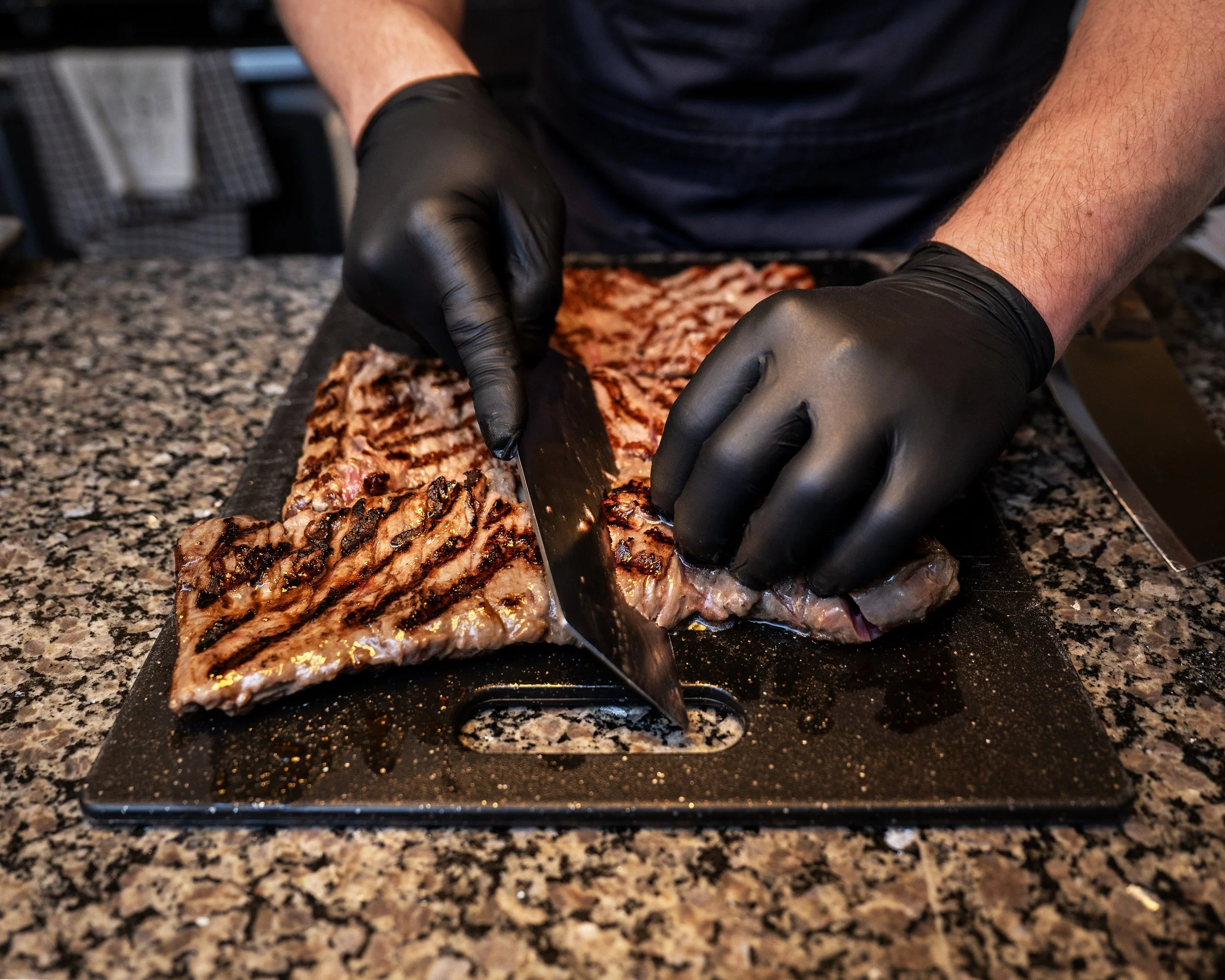 A person wearing black gloves slicing grilled meat on a cutting board in a kitchen with a granite countertop.