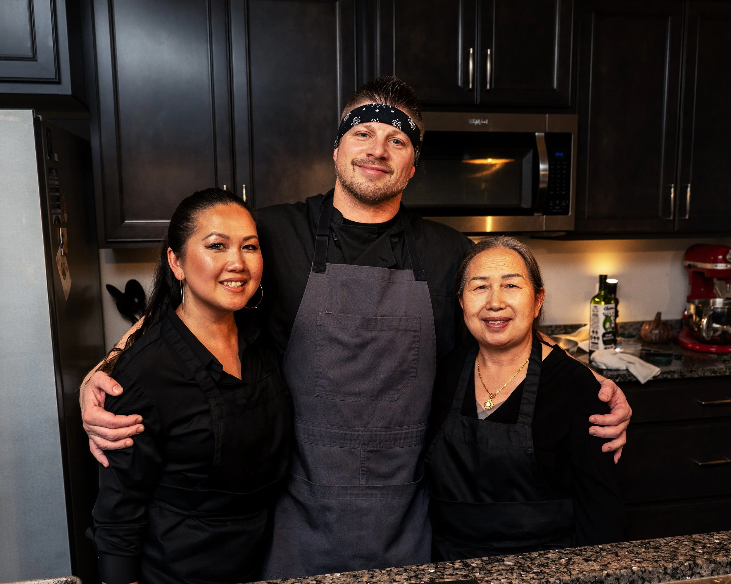 Four people standing together in a kitchen, smiling at the camera. One man is wearing a black bandana and apron, and the three women are also in black tops, with the two on the sides having earrings and necklaces.