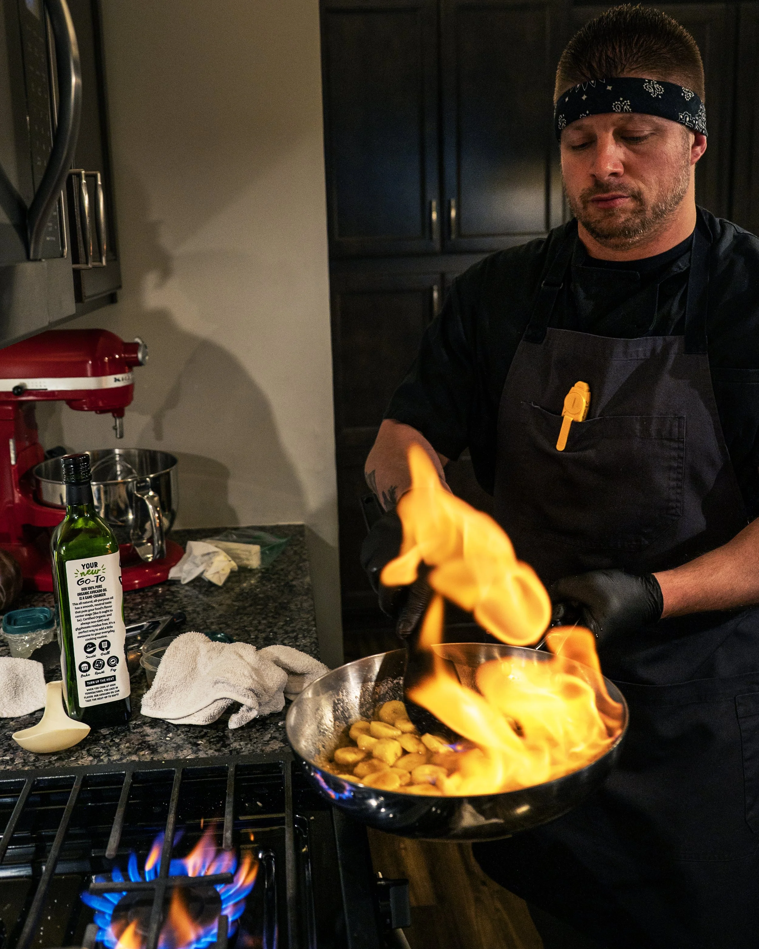 A man wearing a black apron and a black headband is cooking on a stove, holding a pan with flames and food. There are kitchen items like a green olive oil bottle, a red stand mixer, and towels on the counter. Dark cabinets are in the background.