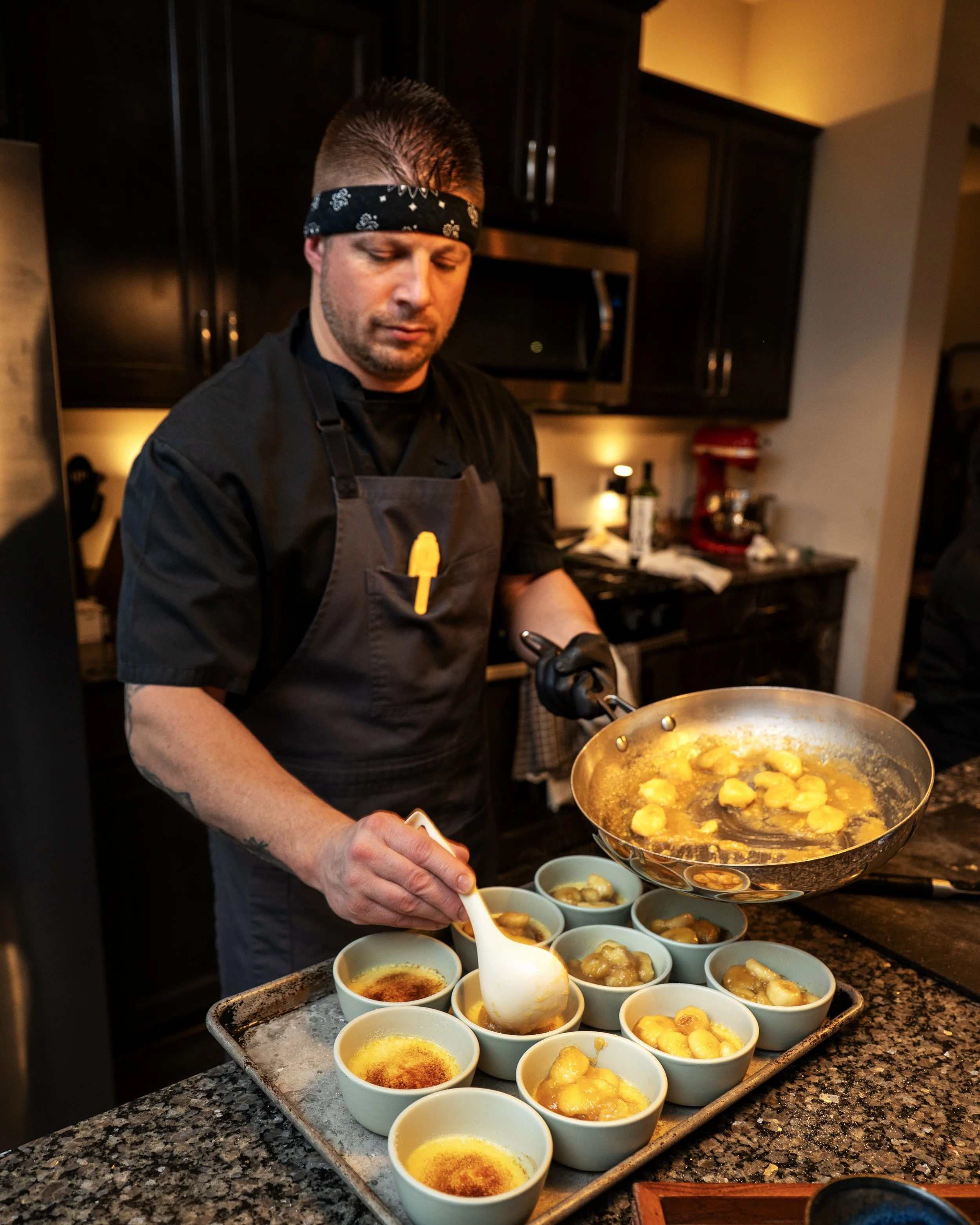 A chef with a bandana on his head is preparing small bowls of dessert in a kitchen. The bowls contain a yellow dessert with a caramelized sugar topping, and some bowls are filled with a creamy, yellow pudding. The chef is holding a ladle and serving 
