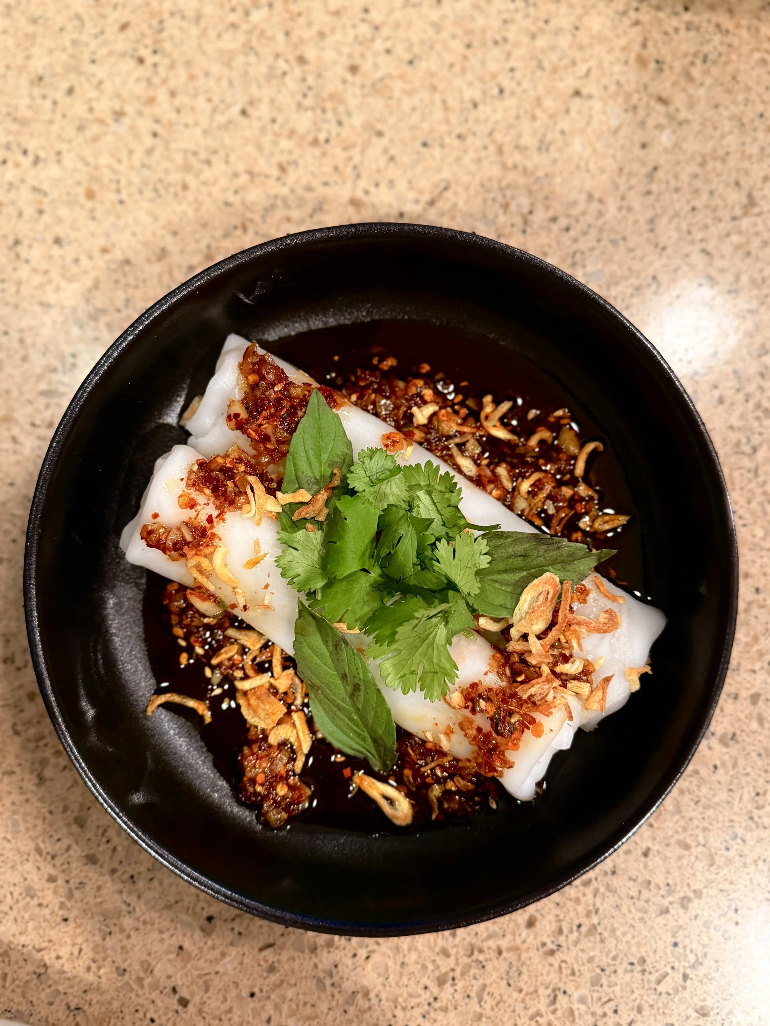 Dumplings topped with chopped garlic, chili flakes, and fresh cilantro, served in a black bowl with soy sauce.